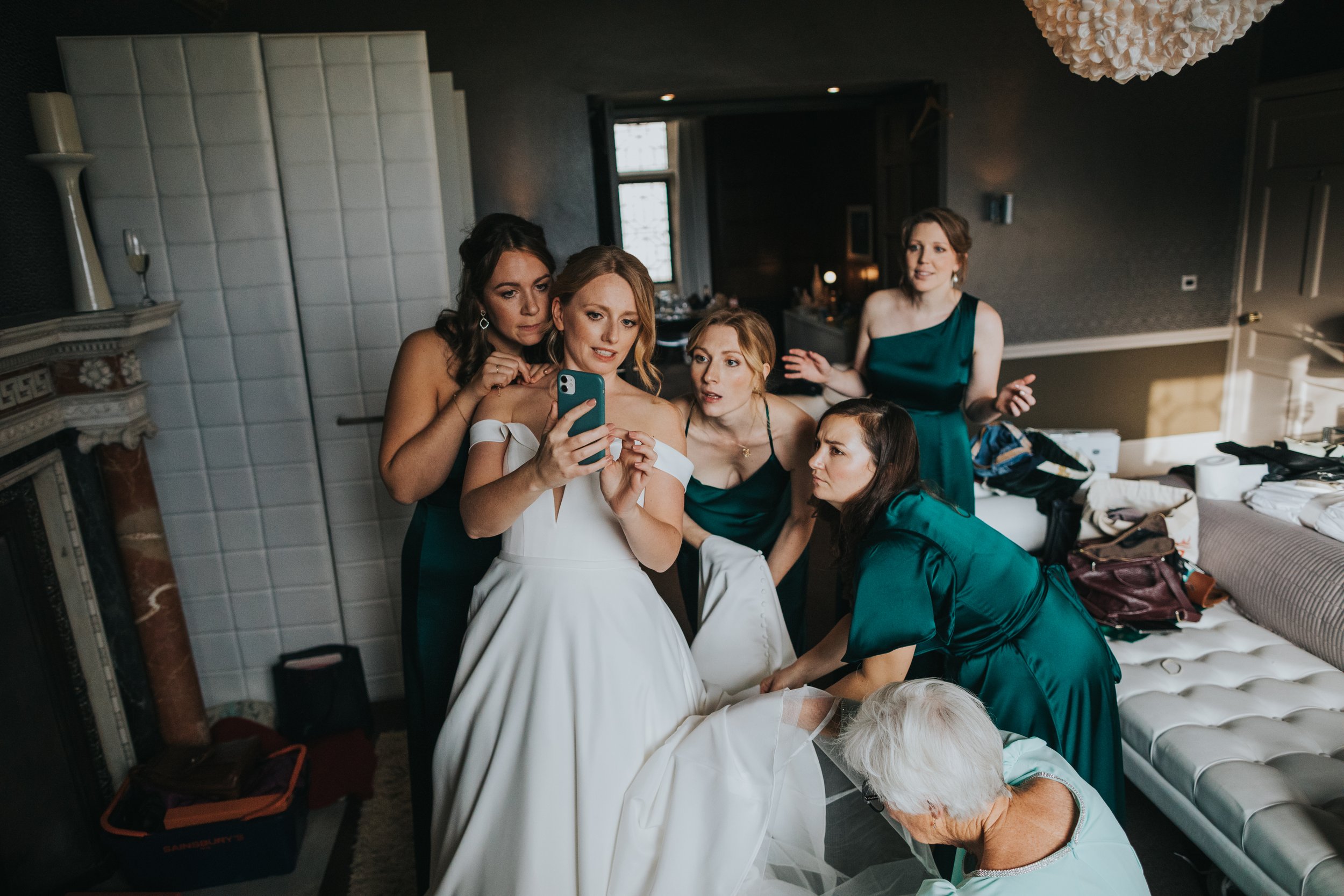 Bridesmaids huddle round to watch the instructions of how to fasten the dress on brides phone. 