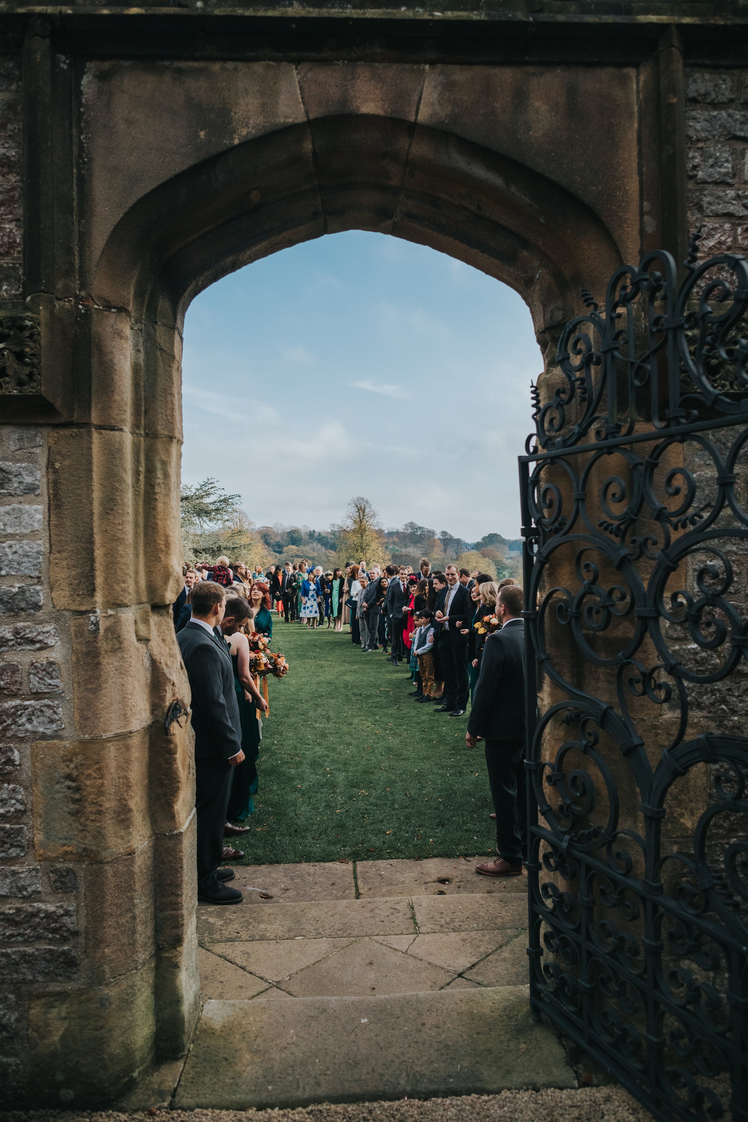 Wedding guests form confetti line through stone arched door with the rolling hills of Derbyshire behind them.