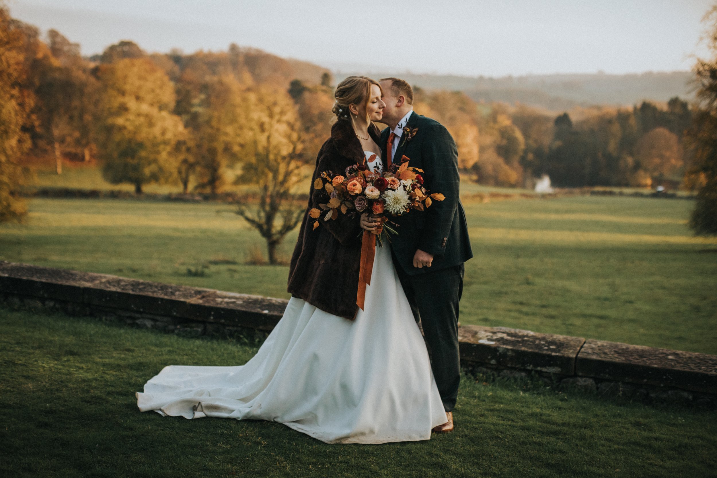 Groom kisses bride in Thornbridge Hall Gardens with the rolling hills of Derbyshire behind them.