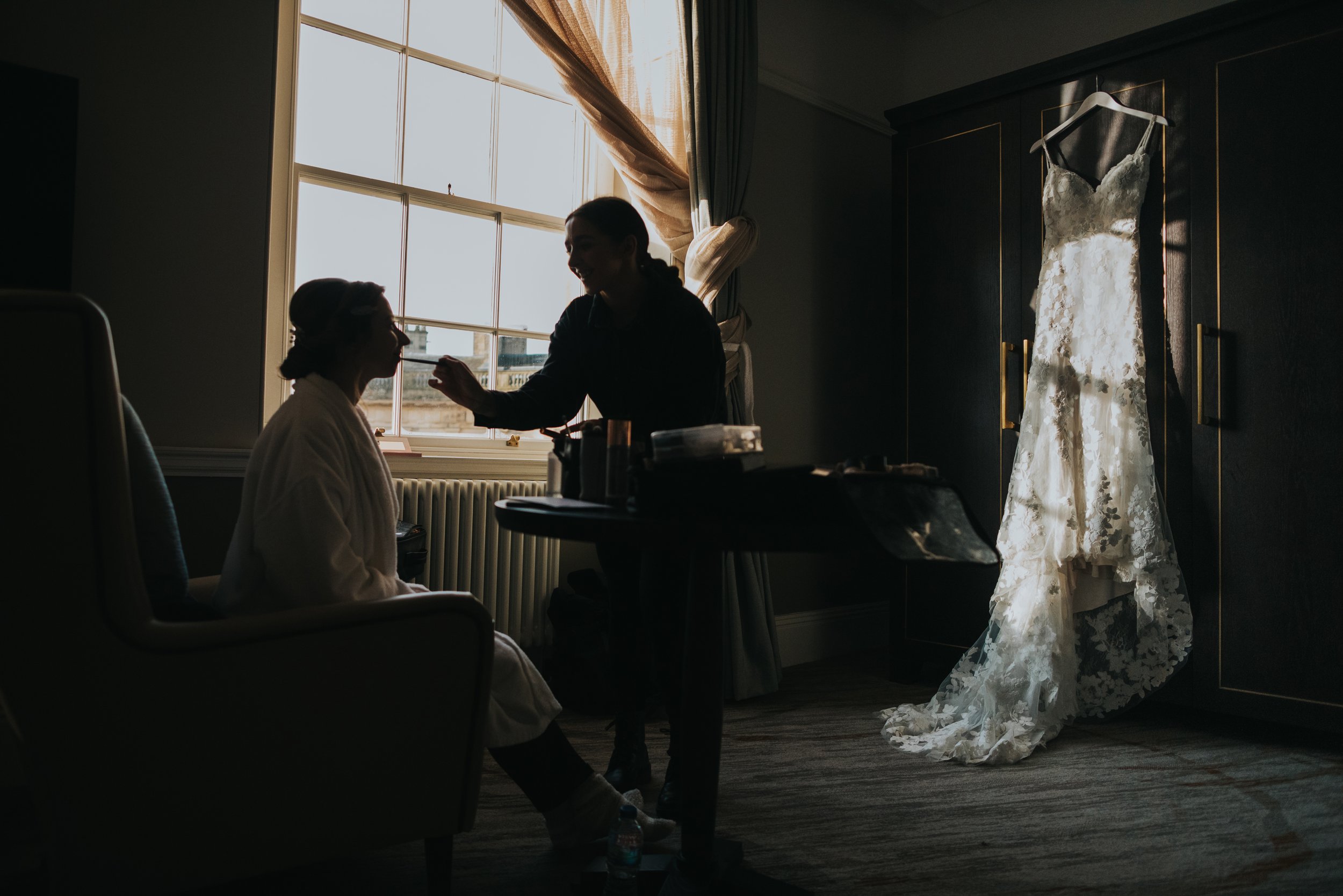 Silhouette of bride having her make up done as her wedding dress hangs up in the back ground. 
