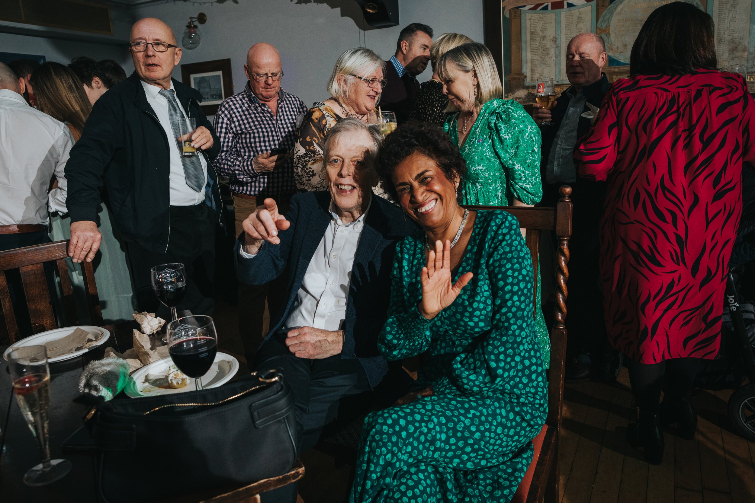 Wedding guests sit at table smiling waving at the camera. 