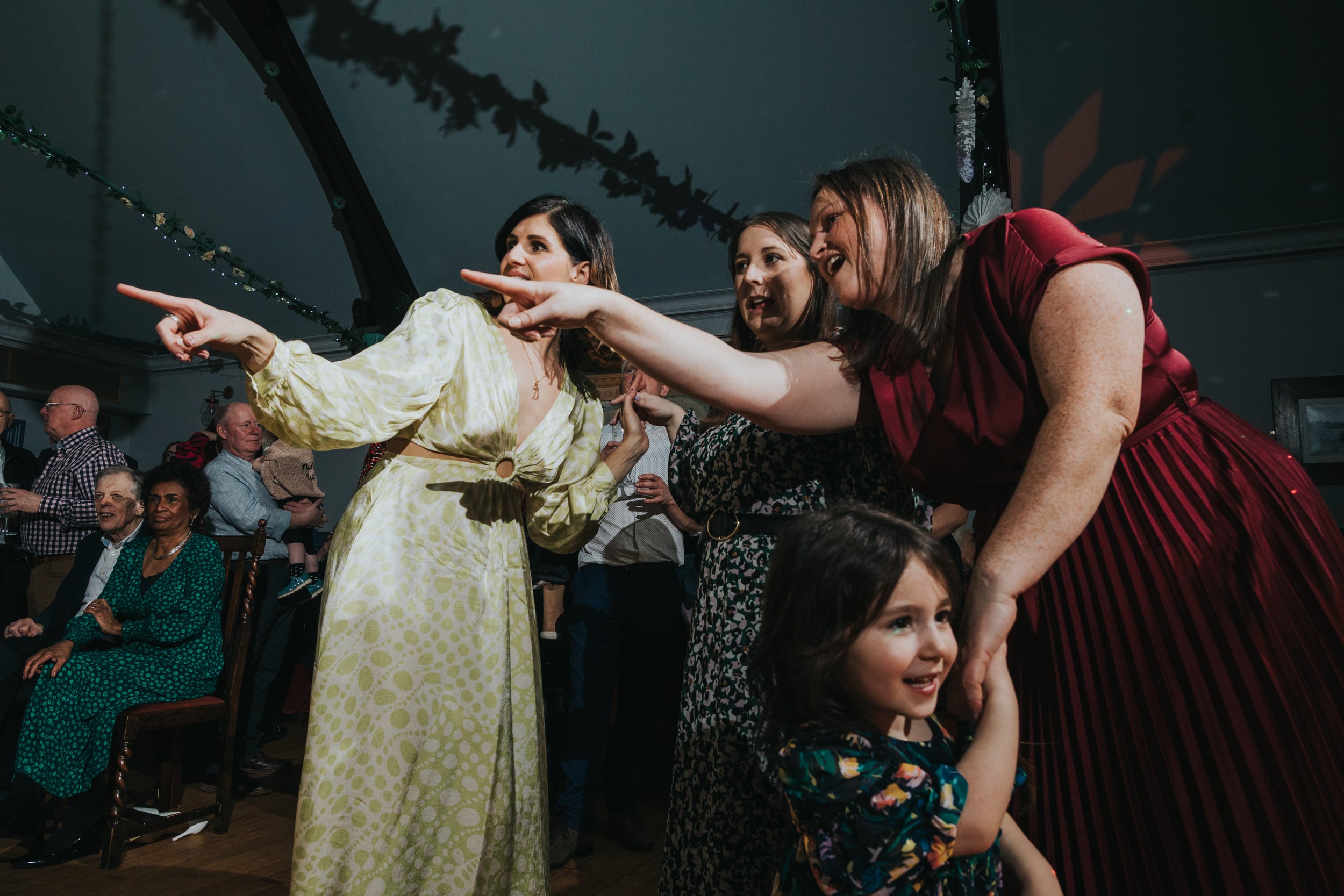 Wedding guests pointing at guest on dance floor as child dances below them. 