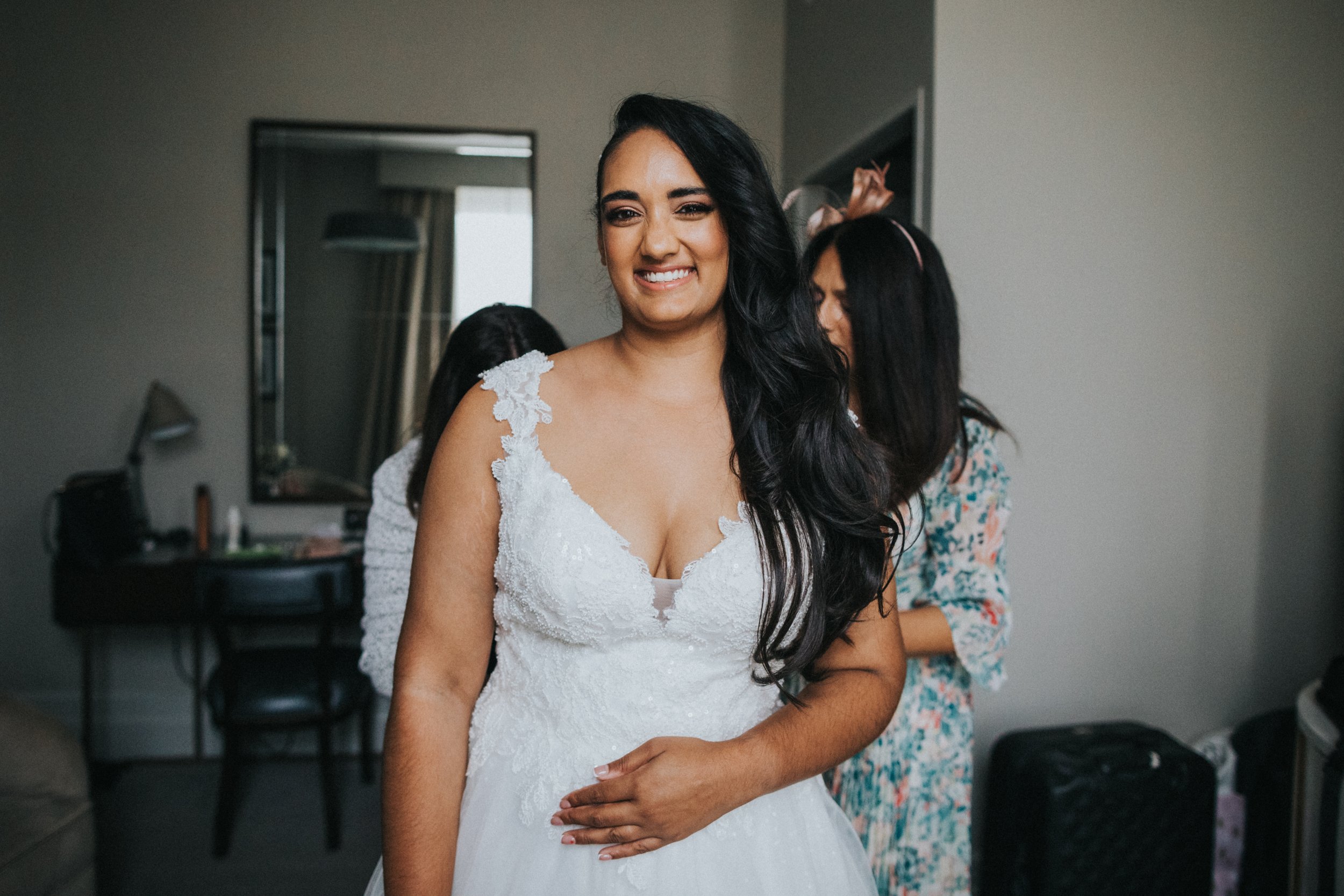 Bride smiles as her mother helps her into her wedding dress. 
