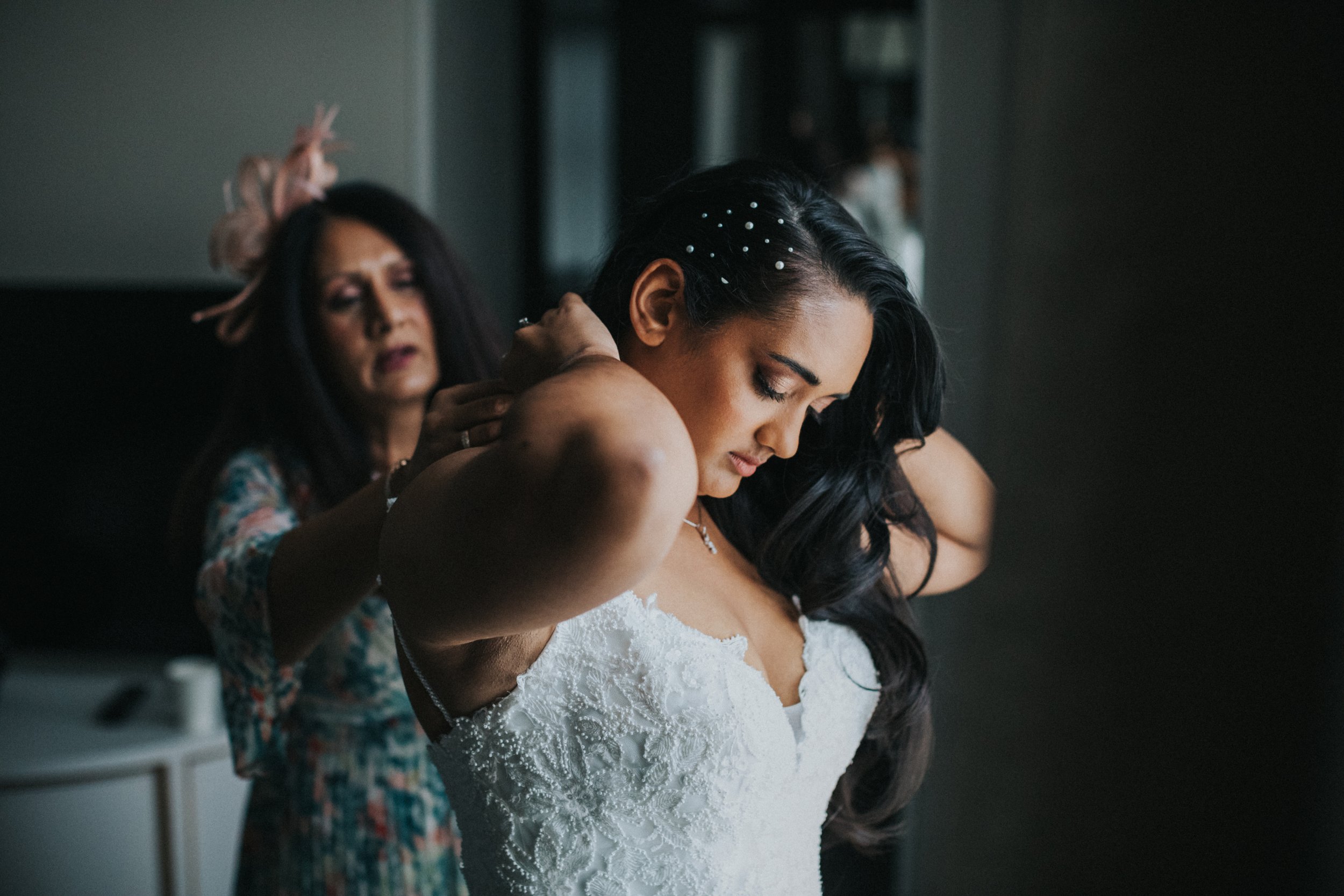 Brides mother helps Bride put on her necklace. 