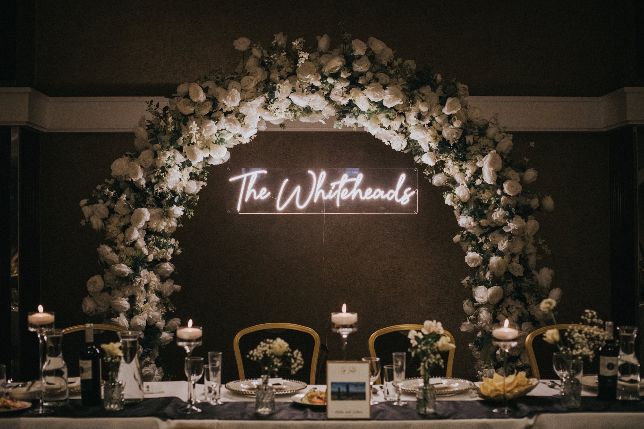 The Whiteheads neon sign underneath a white flower rose arch. 