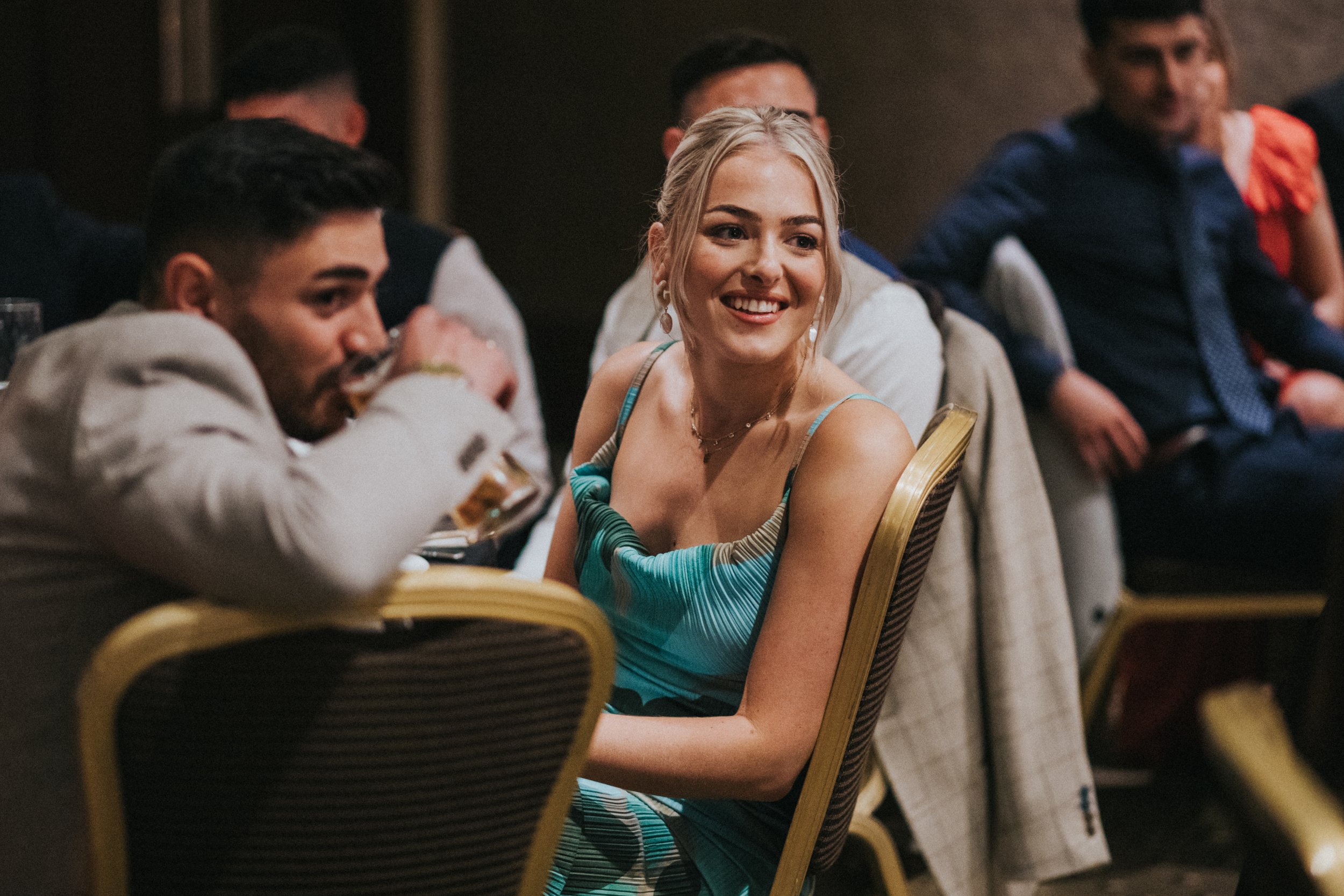 Blonde female wedding guest in blue dress smiles while listening to speeches. 