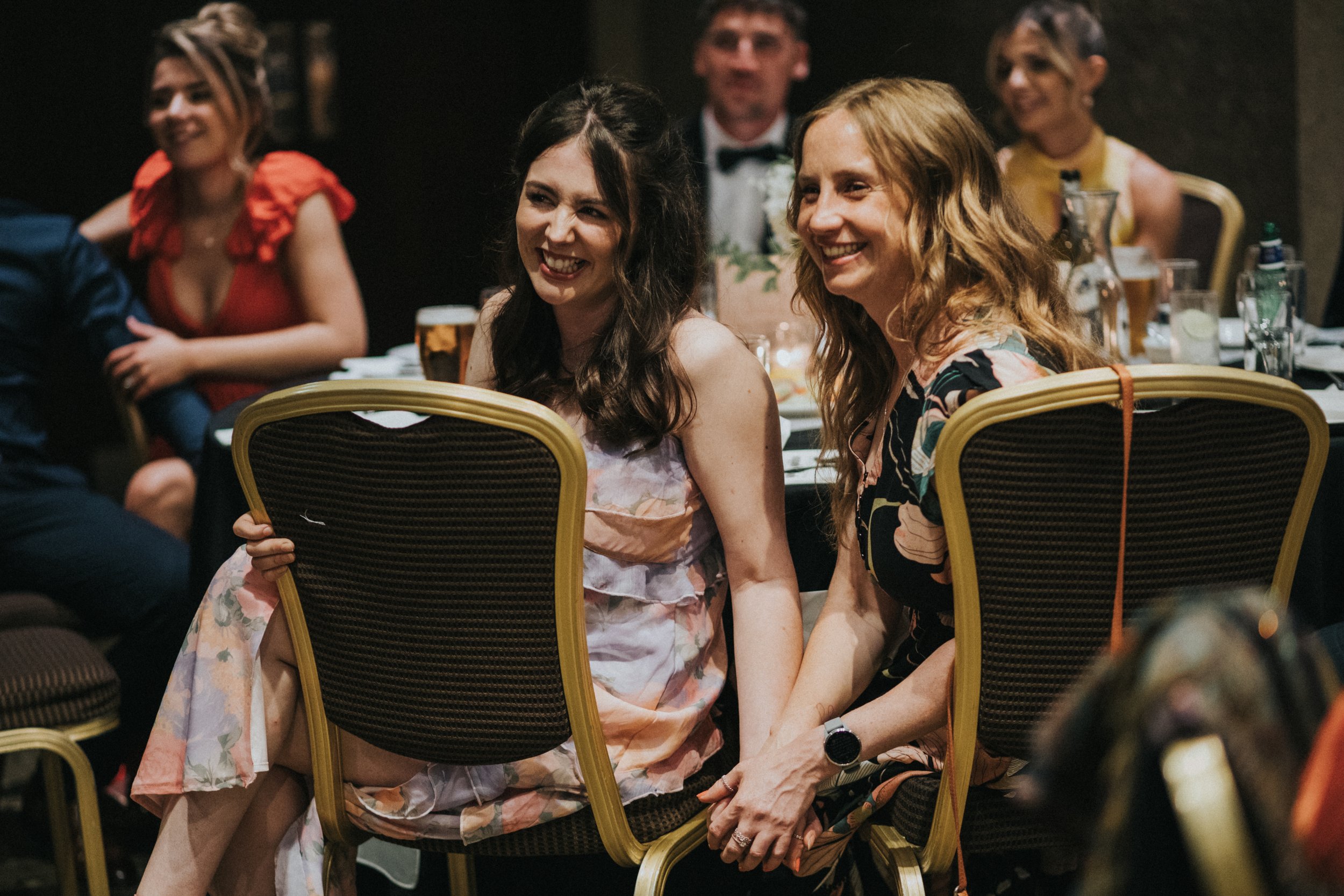 Two female wedding guests hold hands while listening to the speeches. 