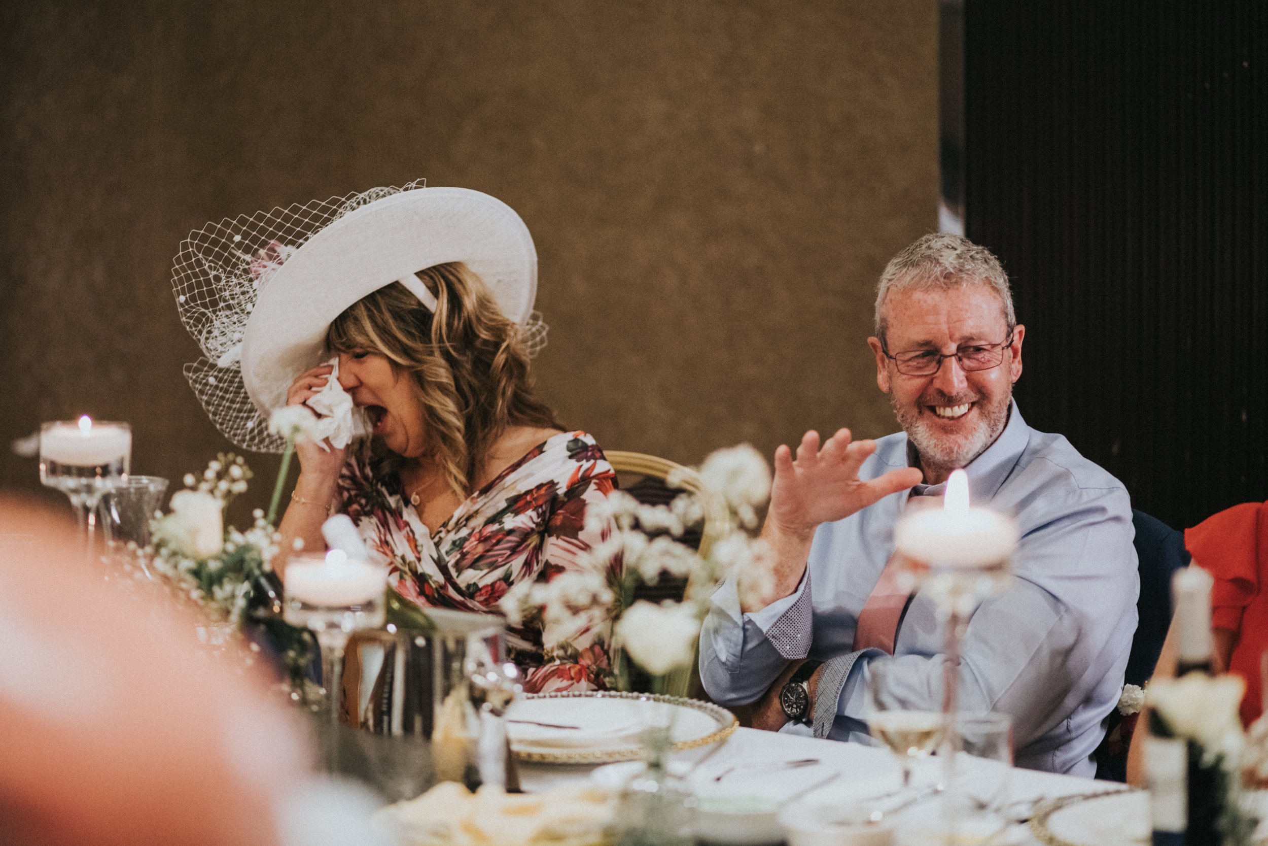 Father of the Groom puts his hand up while his wife sipes tears from her eyes. 