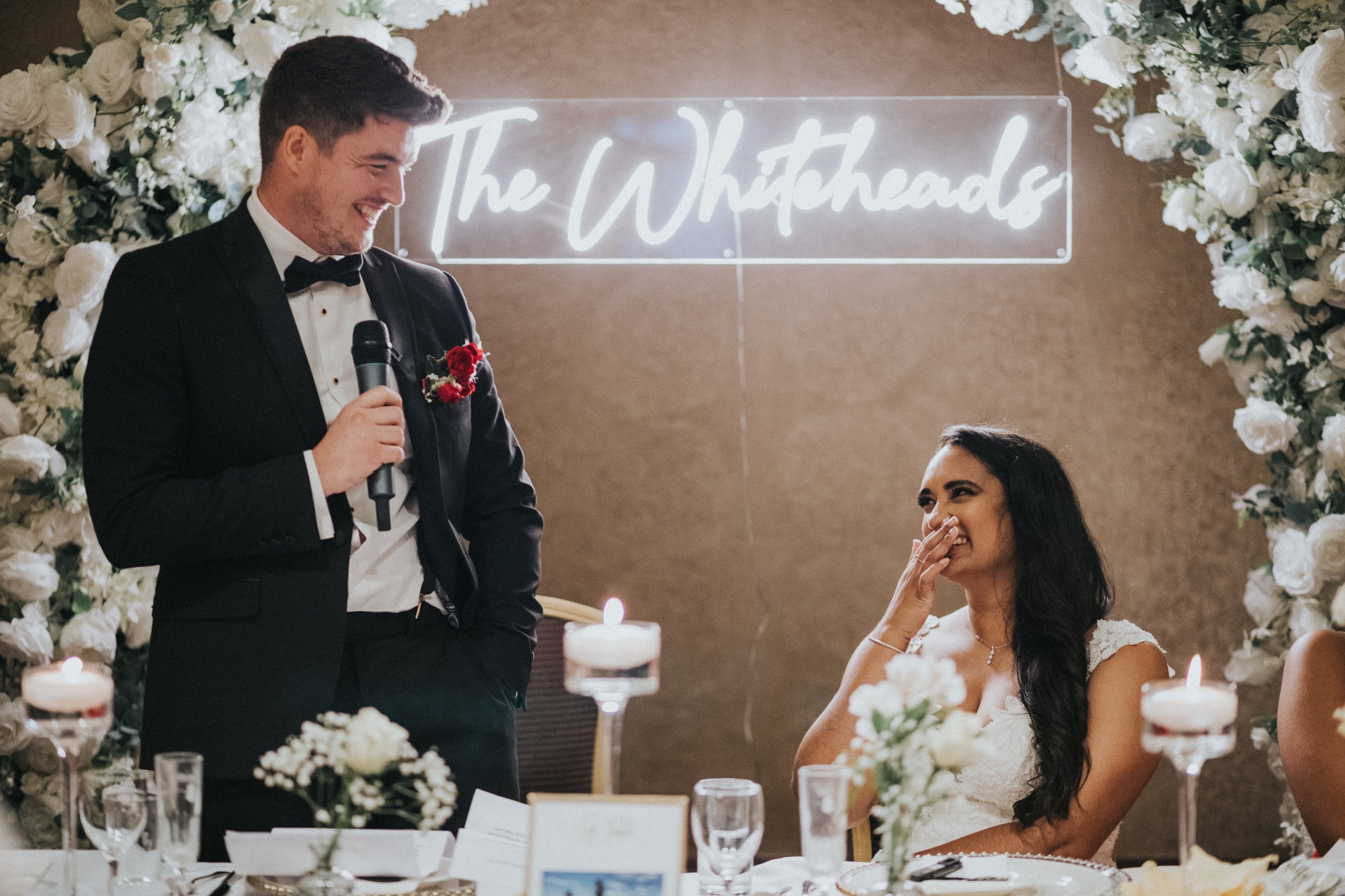 Groom looks at his wife while she laughs at his speech. Whiteheads Neon sign behind them. 