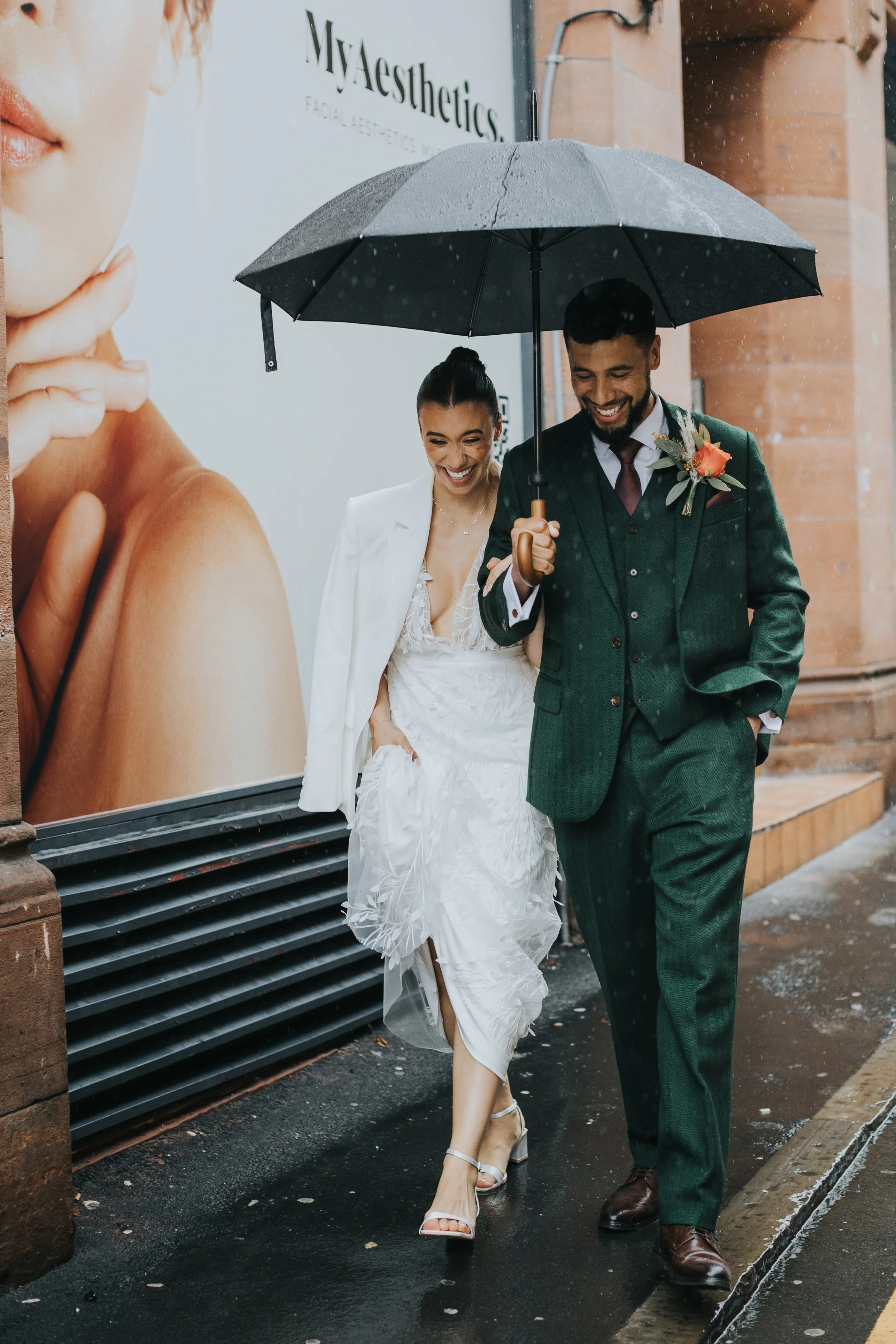 Bride and groom walk down the street laughing under an umbrella on a rainy wedding day in Manchester city centre. 