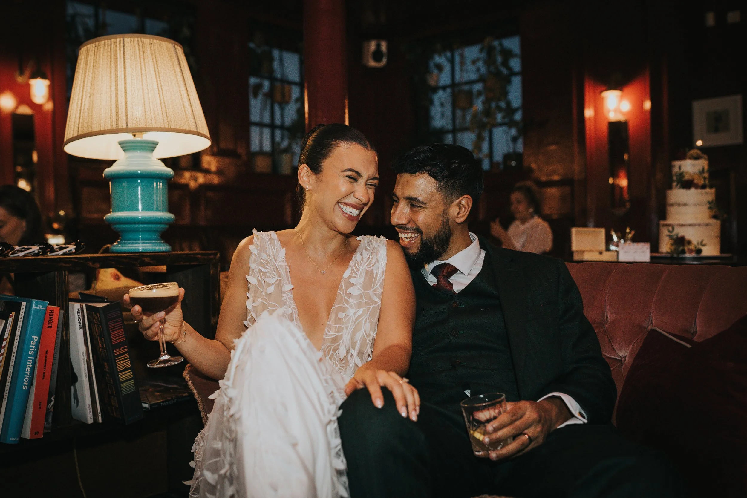 Bride and Groom laugh together in colourful cosy living room like bar setting. 