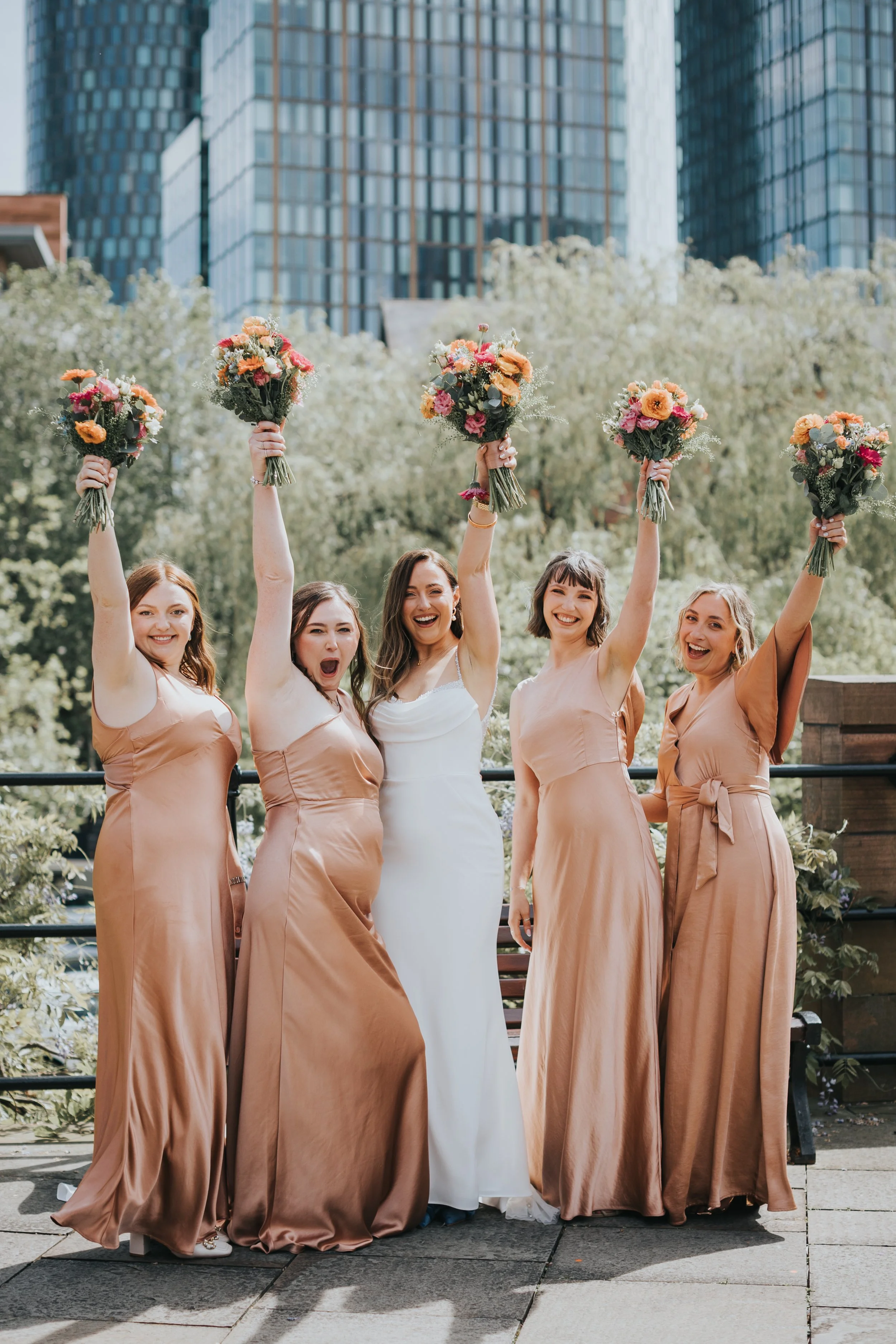Bride and her bridesmaids wearing warm tan dresses holing flowers in the air laughing. 
