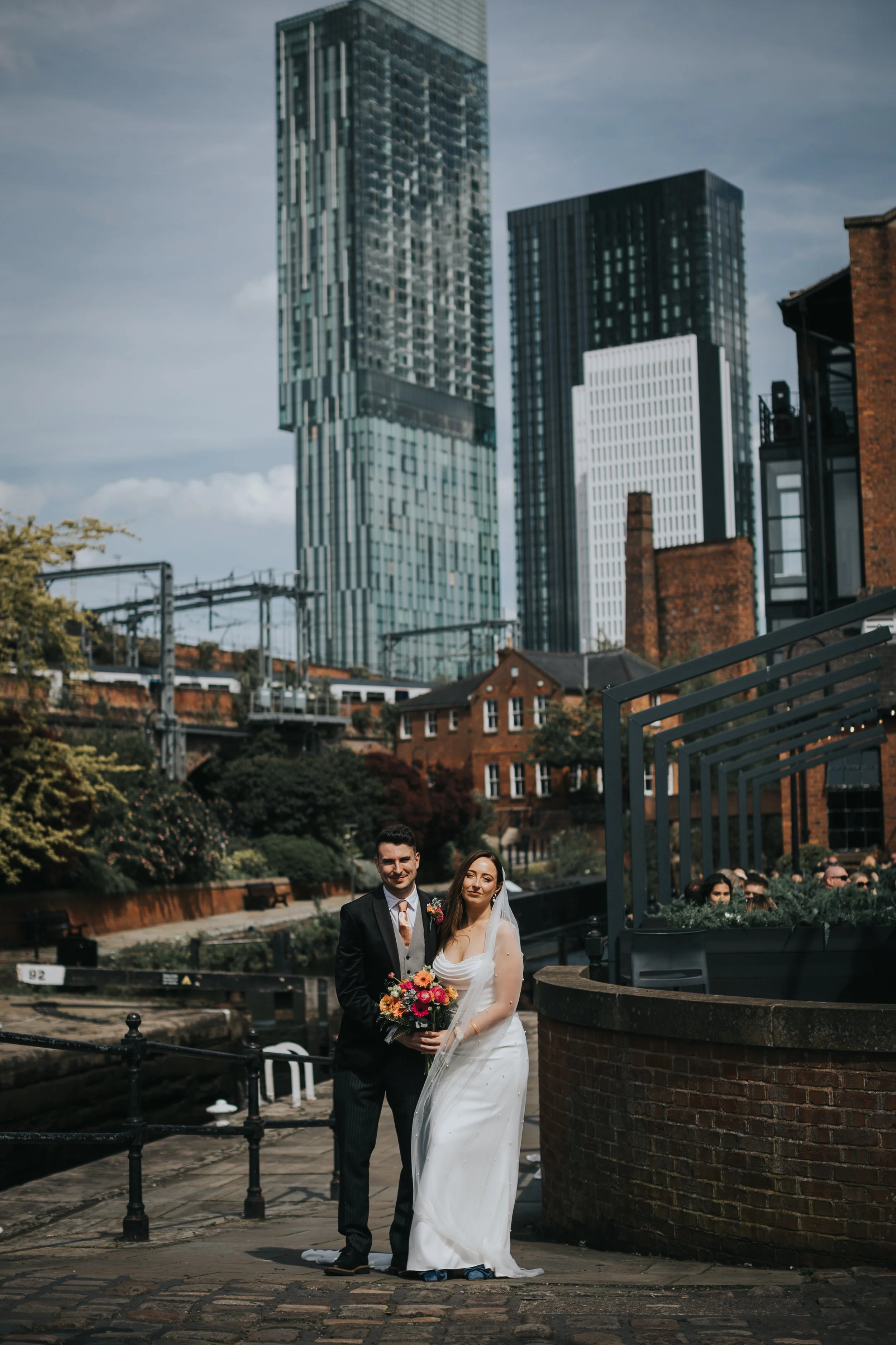 Bride and Groom stand in front of canal lock with sky scrapers behind them. 