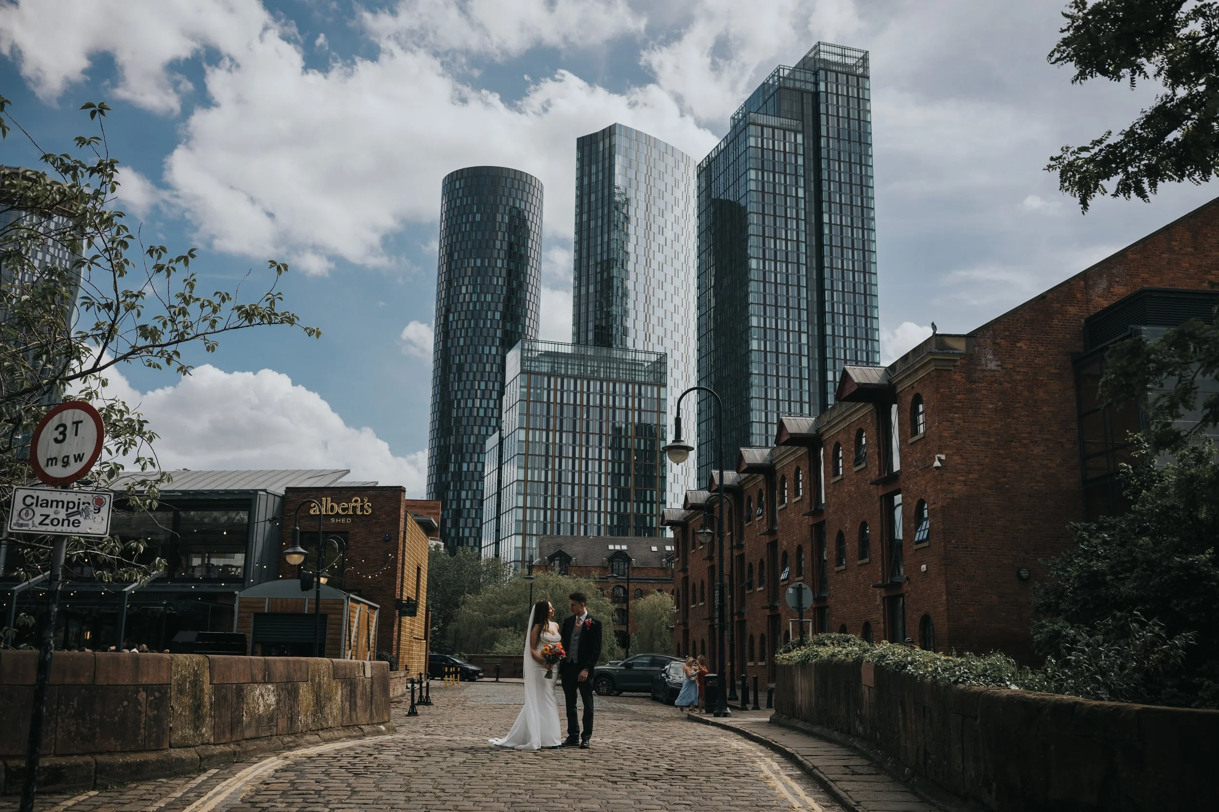 Bride and Groom stand together in the centre of a lock bridge with sky scrapers behind them in Castlefield Manchester.