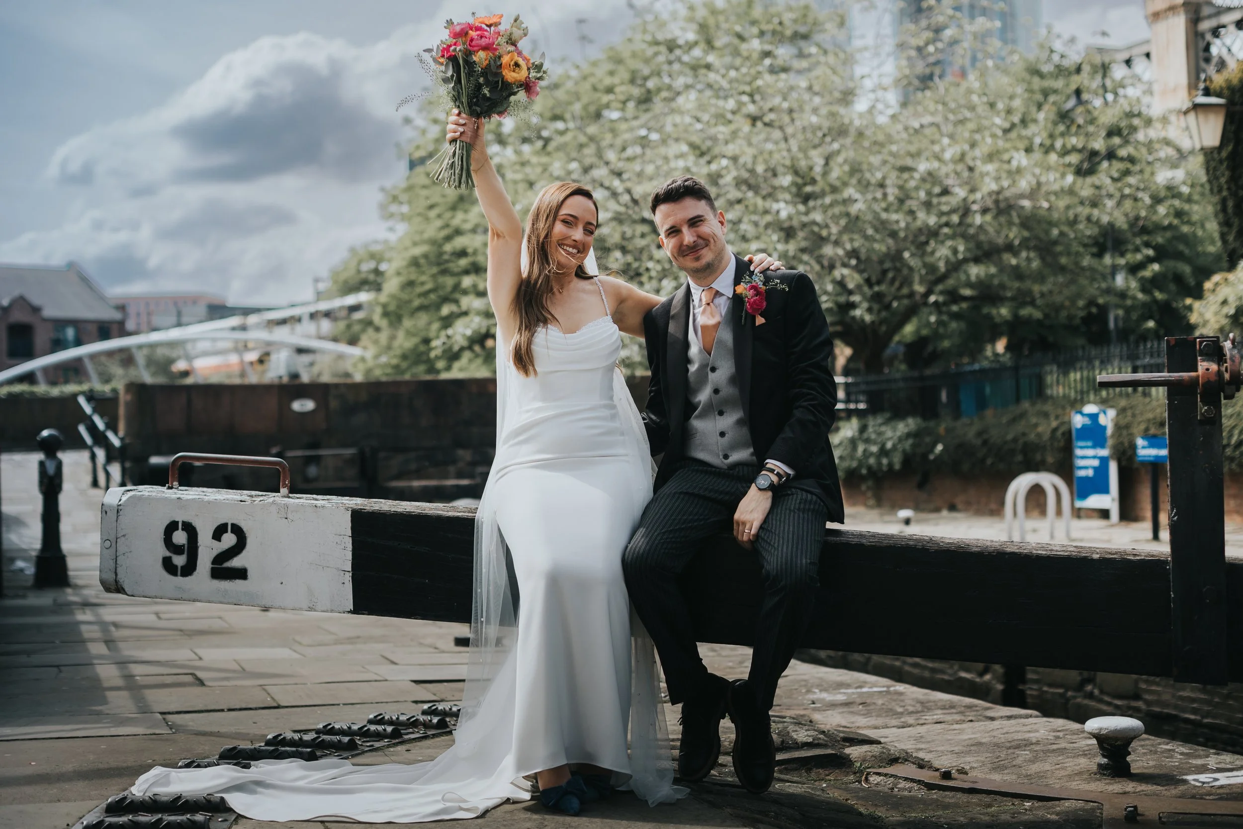 Bride holds flowers up in the air as she sits with her husband on lock in Manchester. 