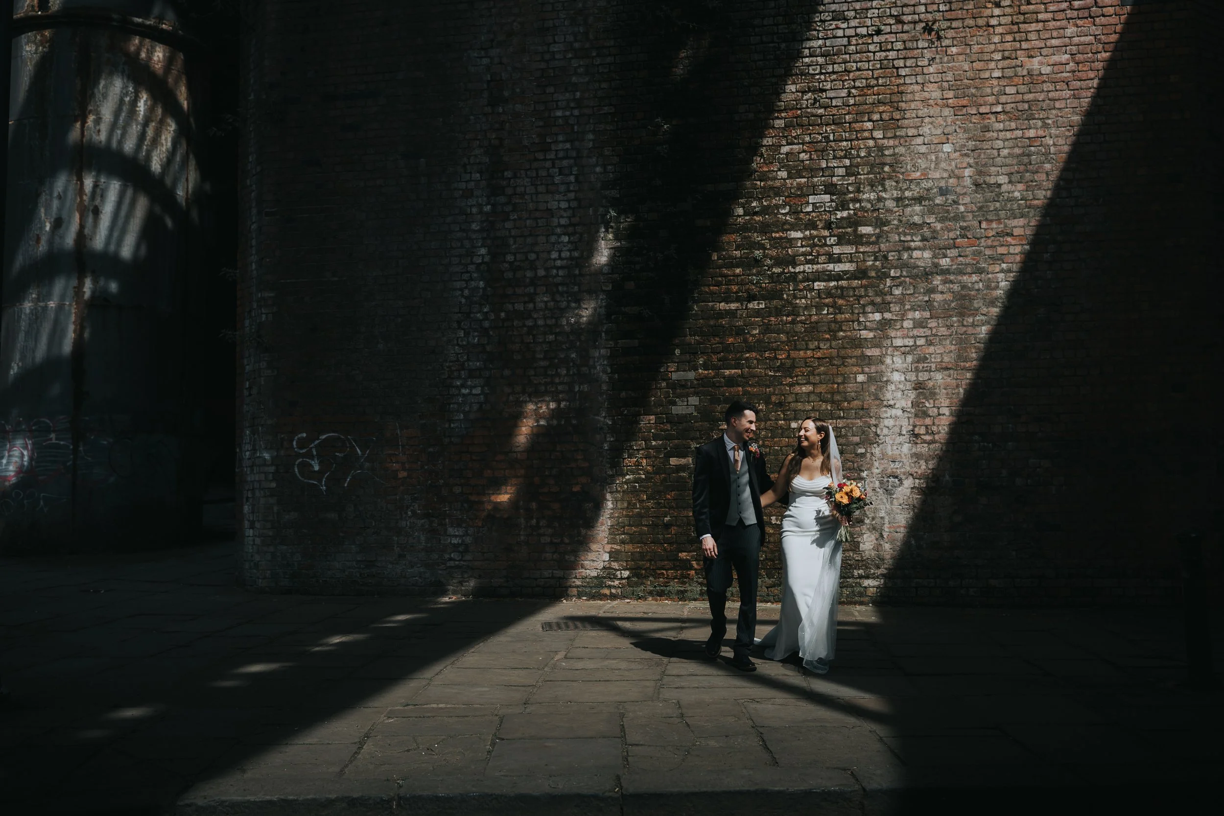 Bride and Groom walk together in dramatic shard of sunlight under Castlefield Arches. 