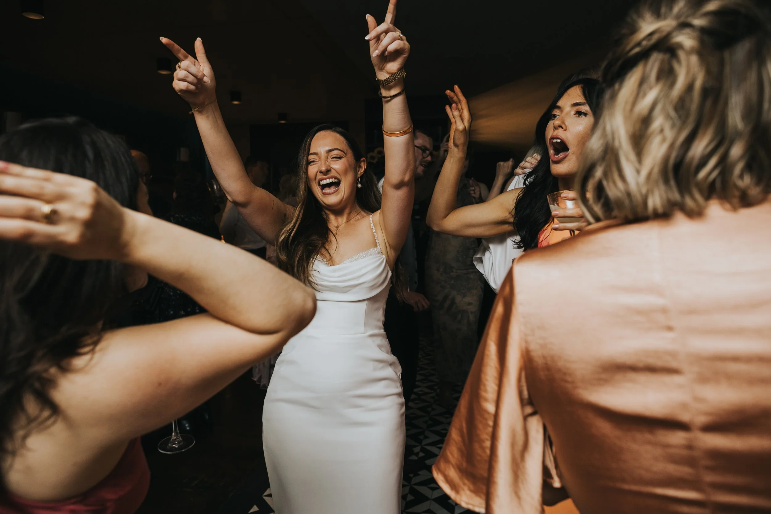 Bride has her hands in the air laughing as she dances with her bridesmaids. 