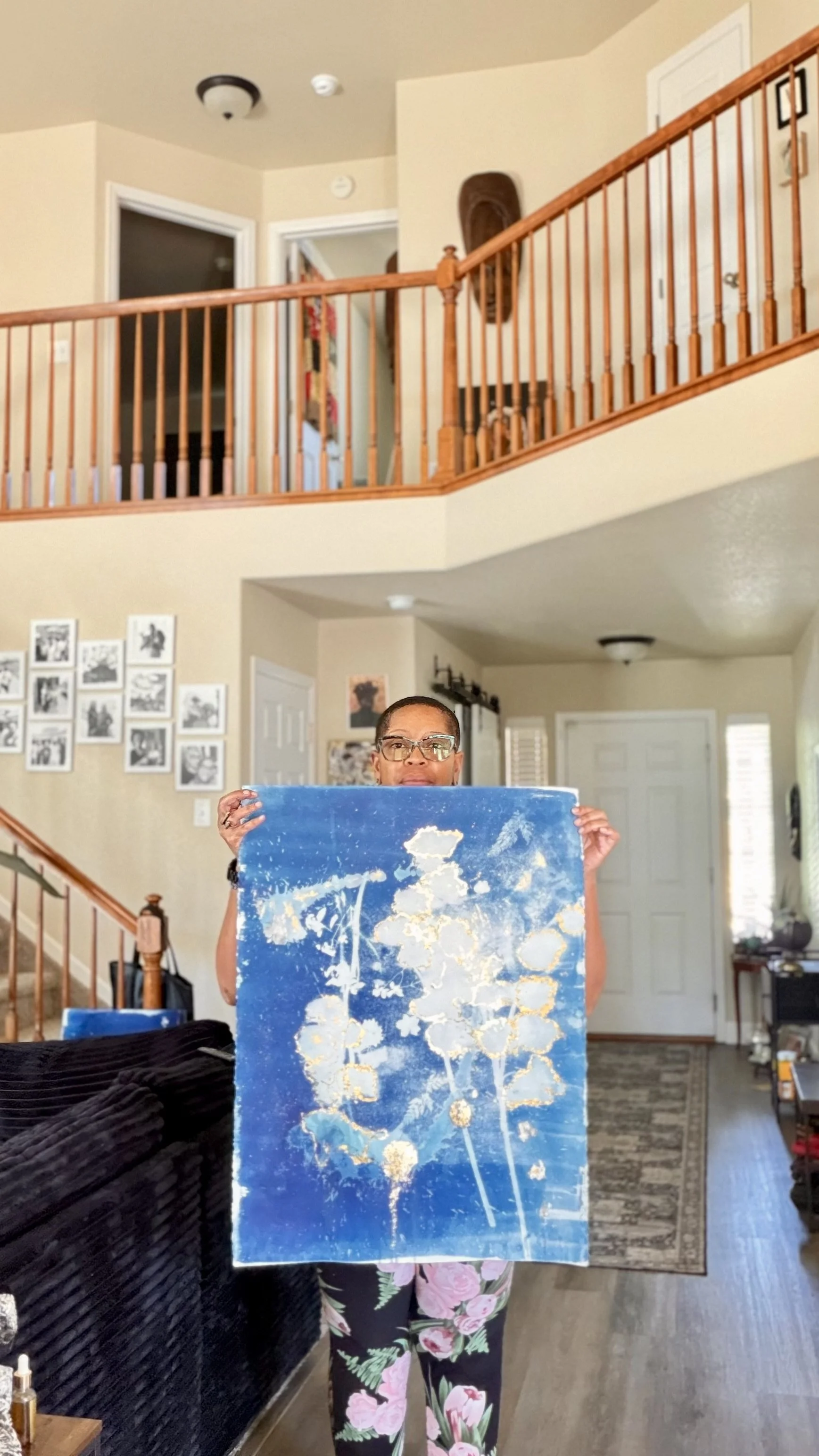 A woman in glasses and floral pants holding an abstract blue and gold painting in a living room.
