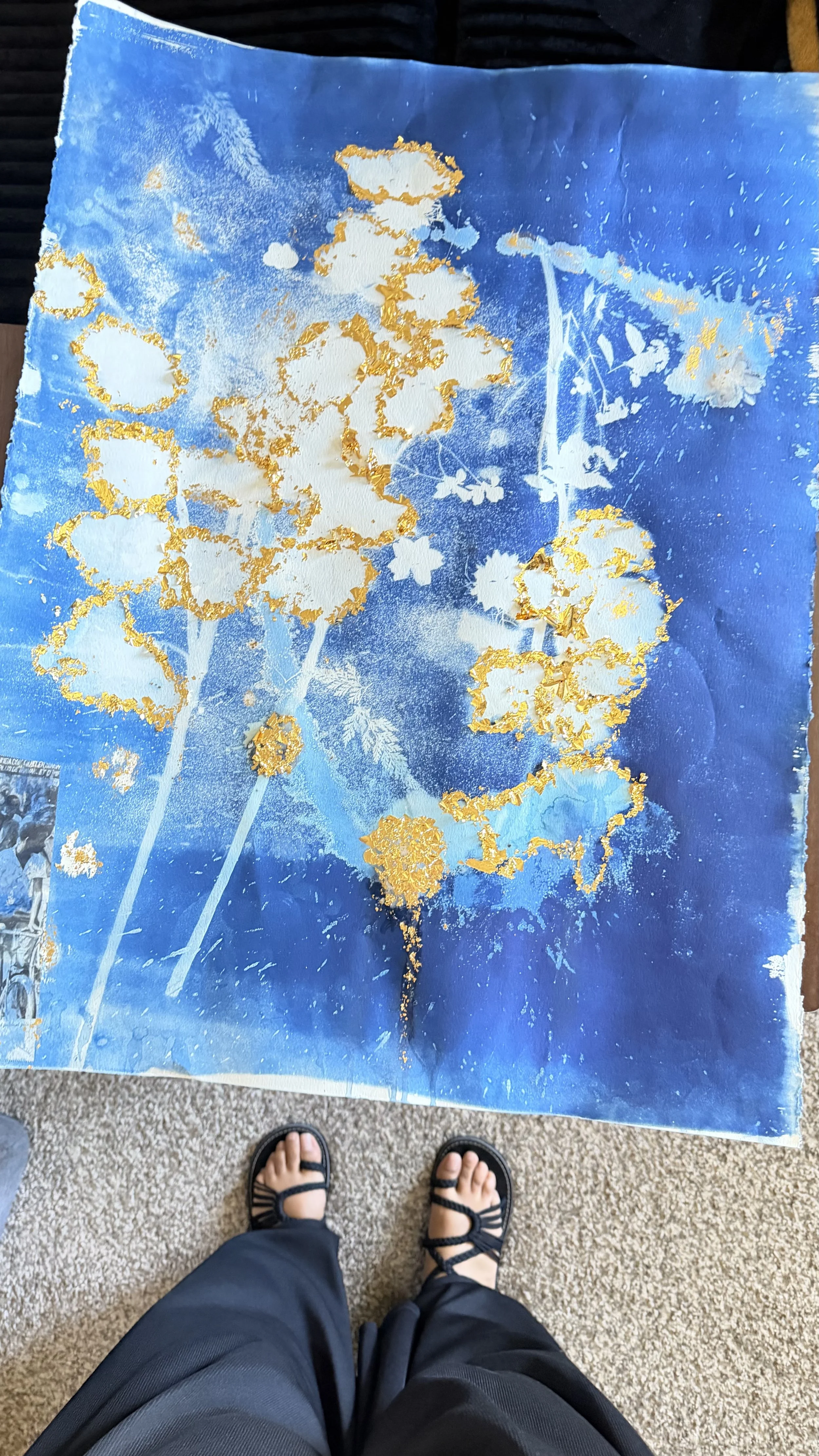 Person standing on a beige carpeted floor wearing black sandals and dark pants, with a colorful paper craft art project laid flat on the floor. The art features blue, white, and gold colors with floral and abstract patterns.