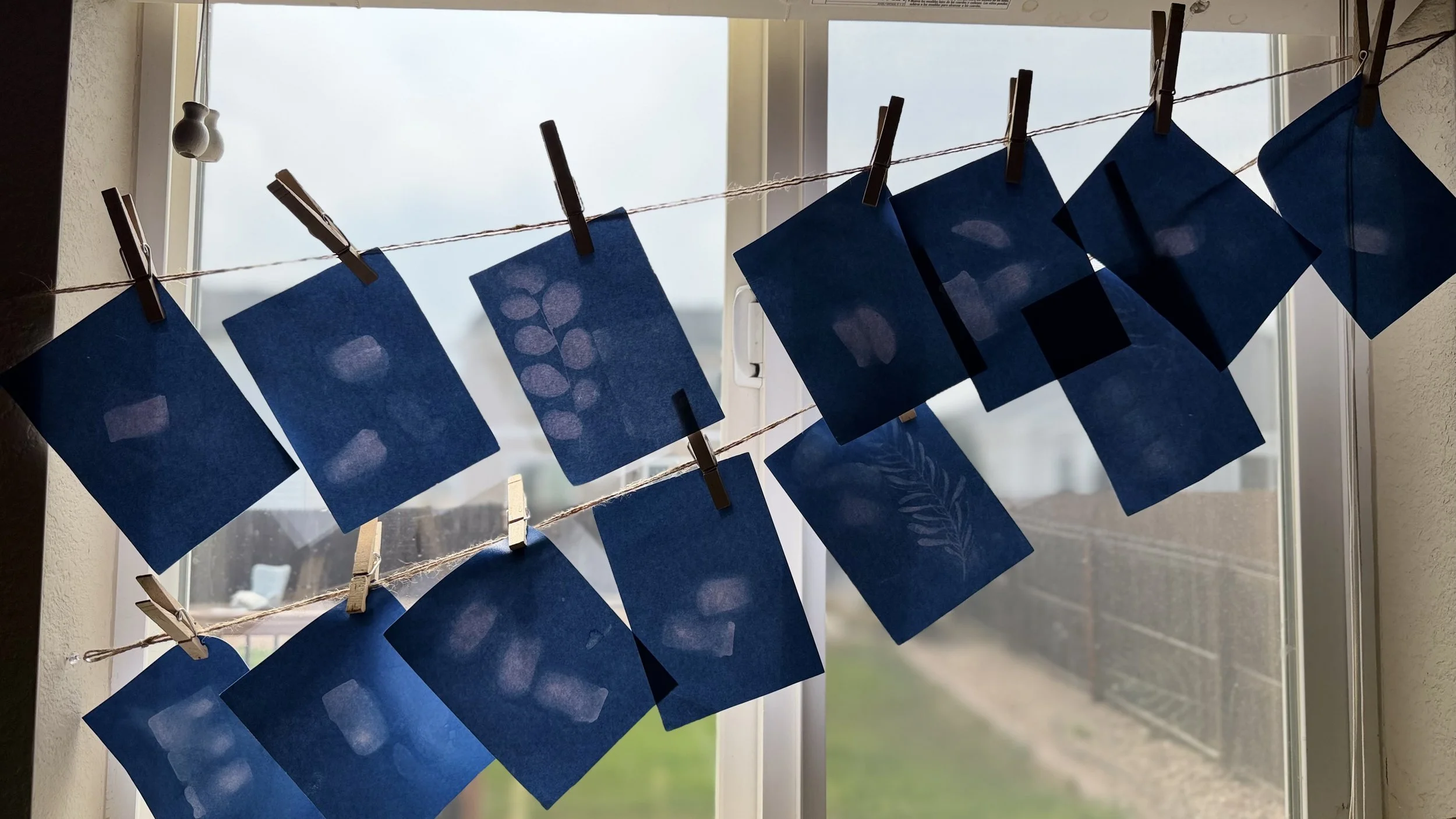 Blue paper cards with leaf and plant designs hanging on a string outside near a window, secured with clothespins, with a cloudy sky in the background.
