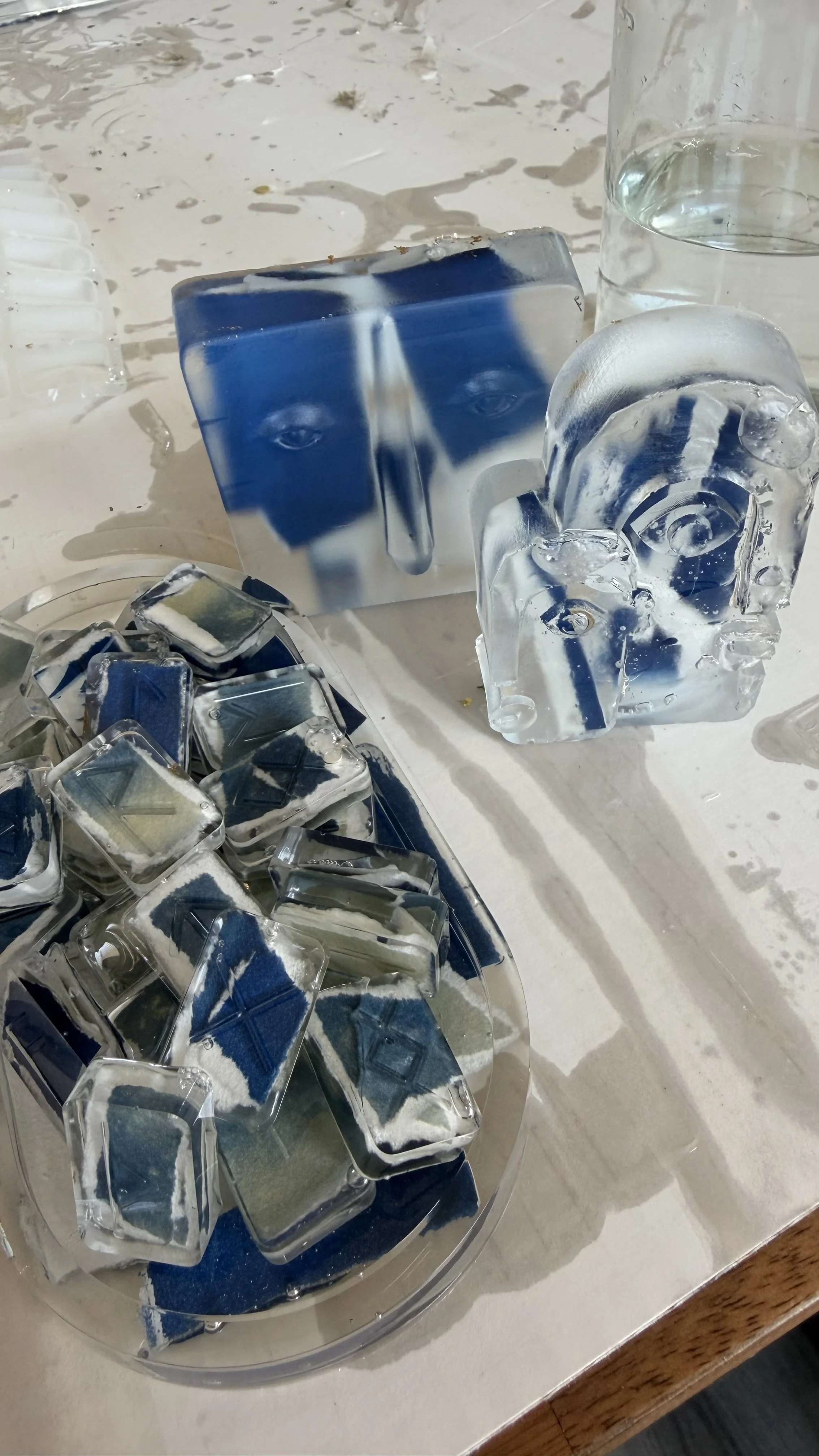 Ice cubes with a blue tinge, some shaped like human faces, arranged on a table with a glass of water nearby.
