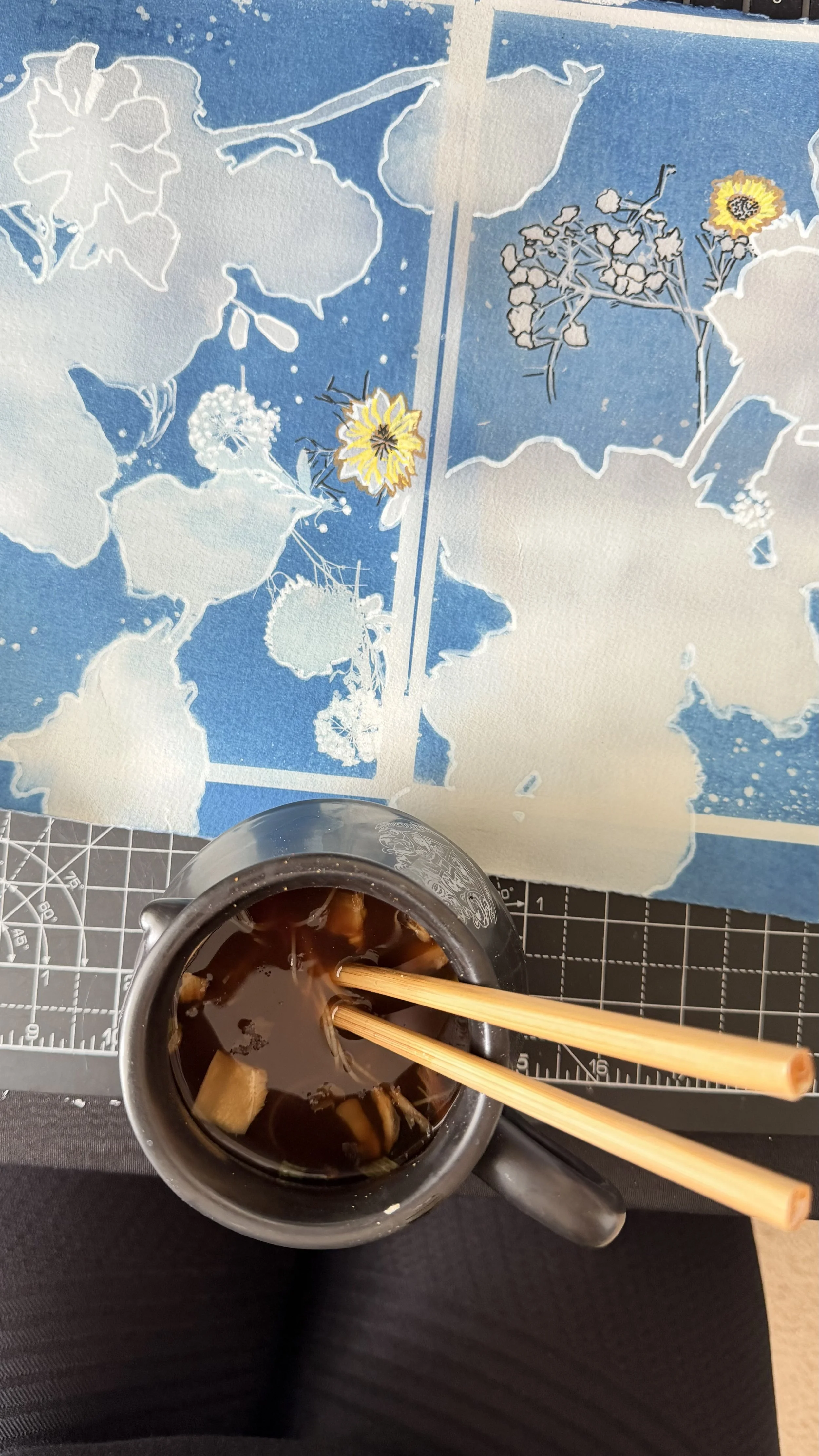 A menu with a blue floral pattern background, a pair of chopsticks resting over a small bowl of dark soup with ingredients, and a person's black pants visible at the bottom.