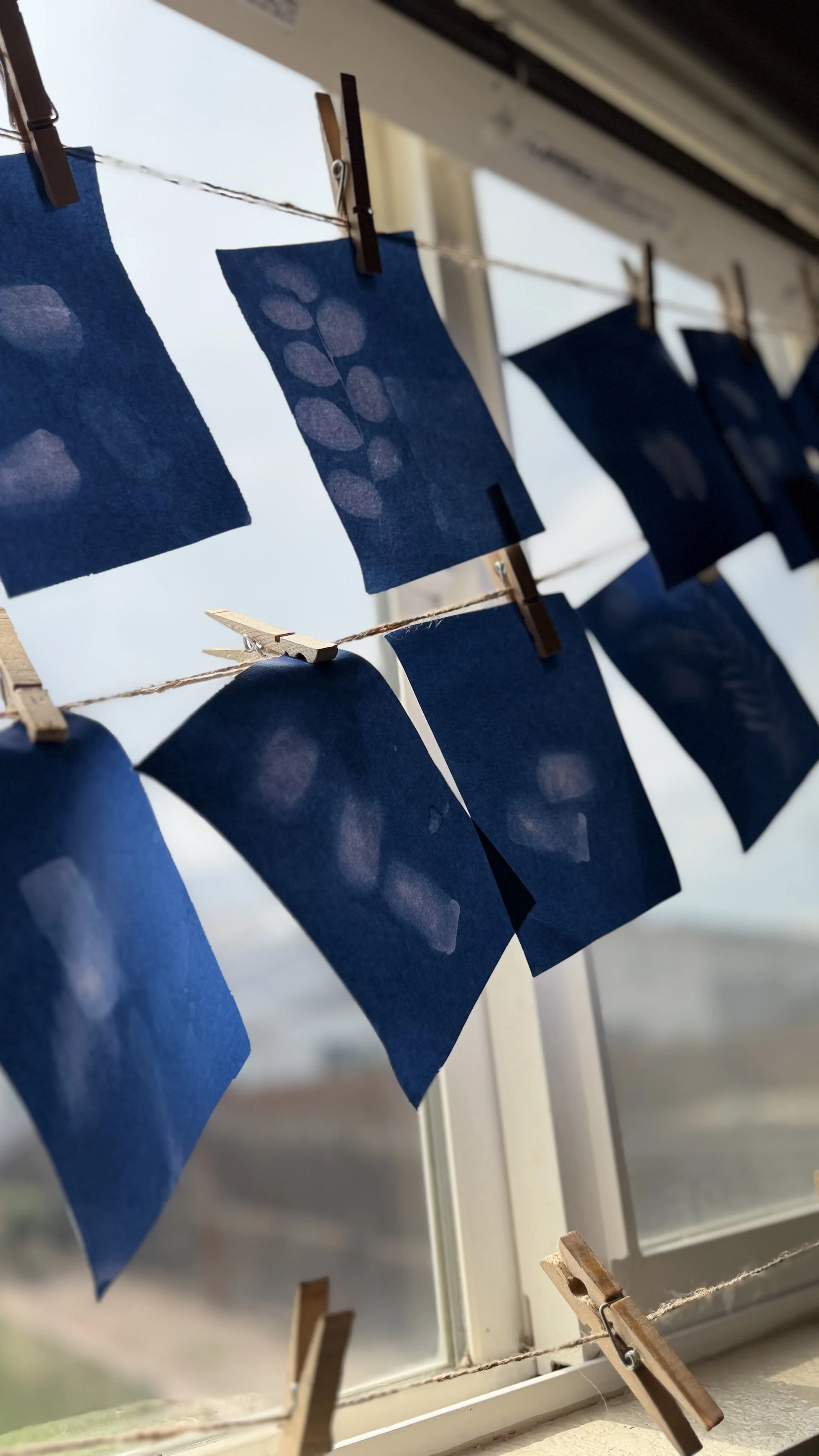 Blue paper with leaf patterns hanging on a clothesline with clothespins near a window.