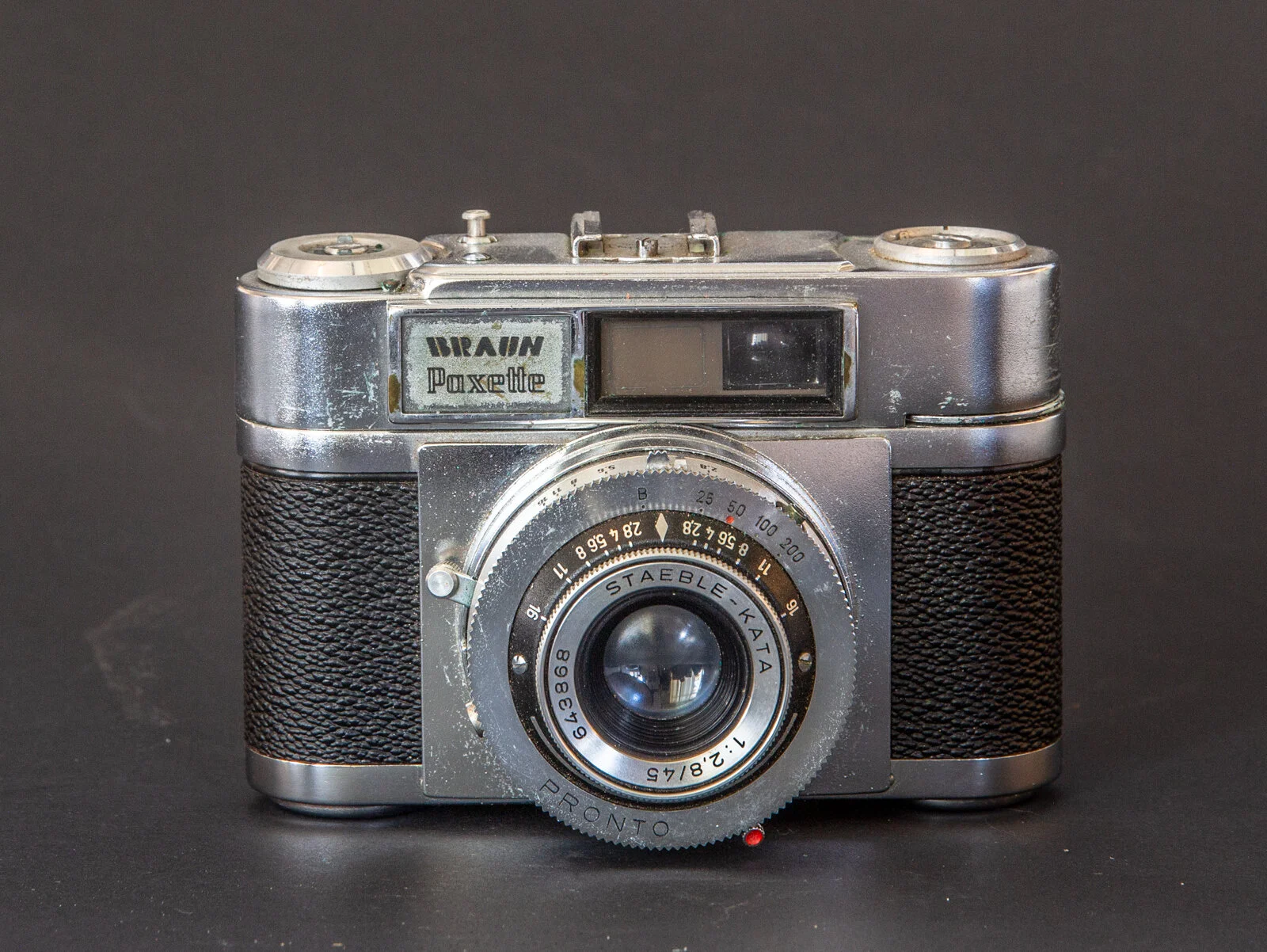 Ancienne caméra argentée avec grip noir, objectif rond marqué Staeble-Kata, cadran, et vue du viseur, sur fond noir.