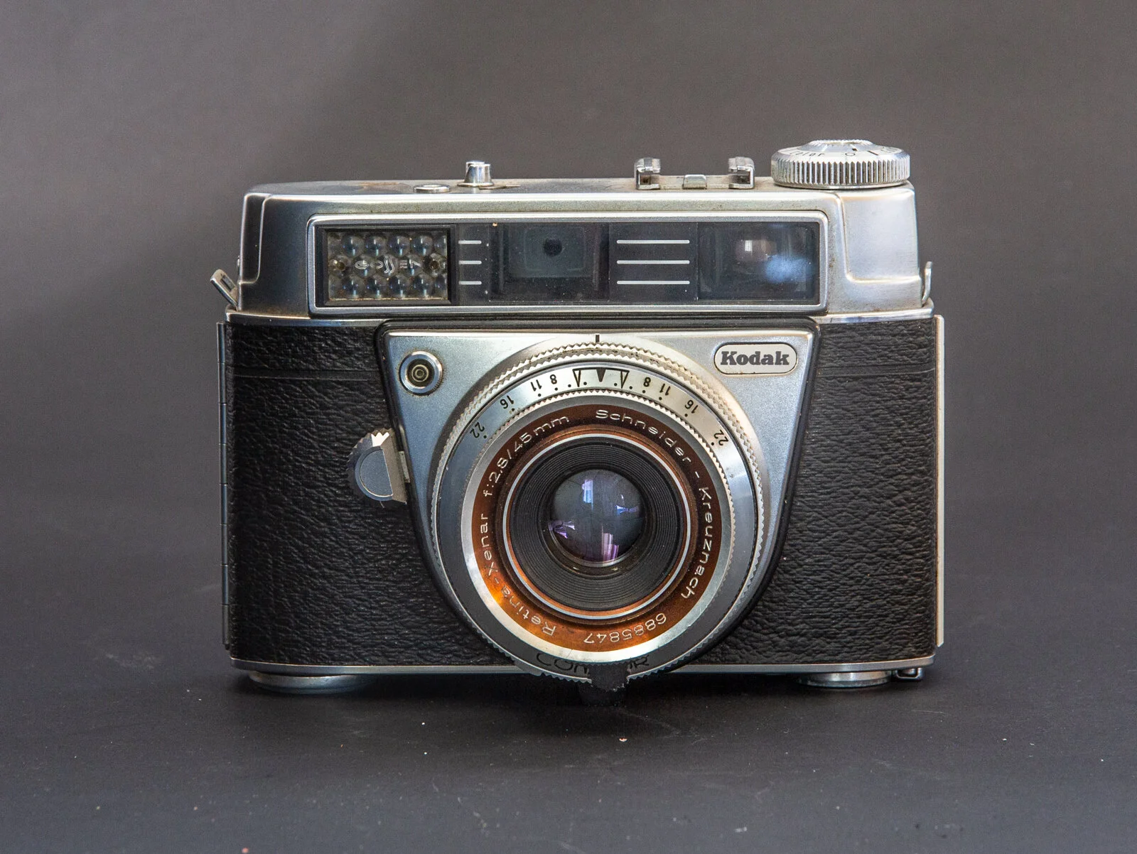 Une caméra vintage Kodak avec objectif en métal et boîtier en cuir noir, placé sur un fond gris