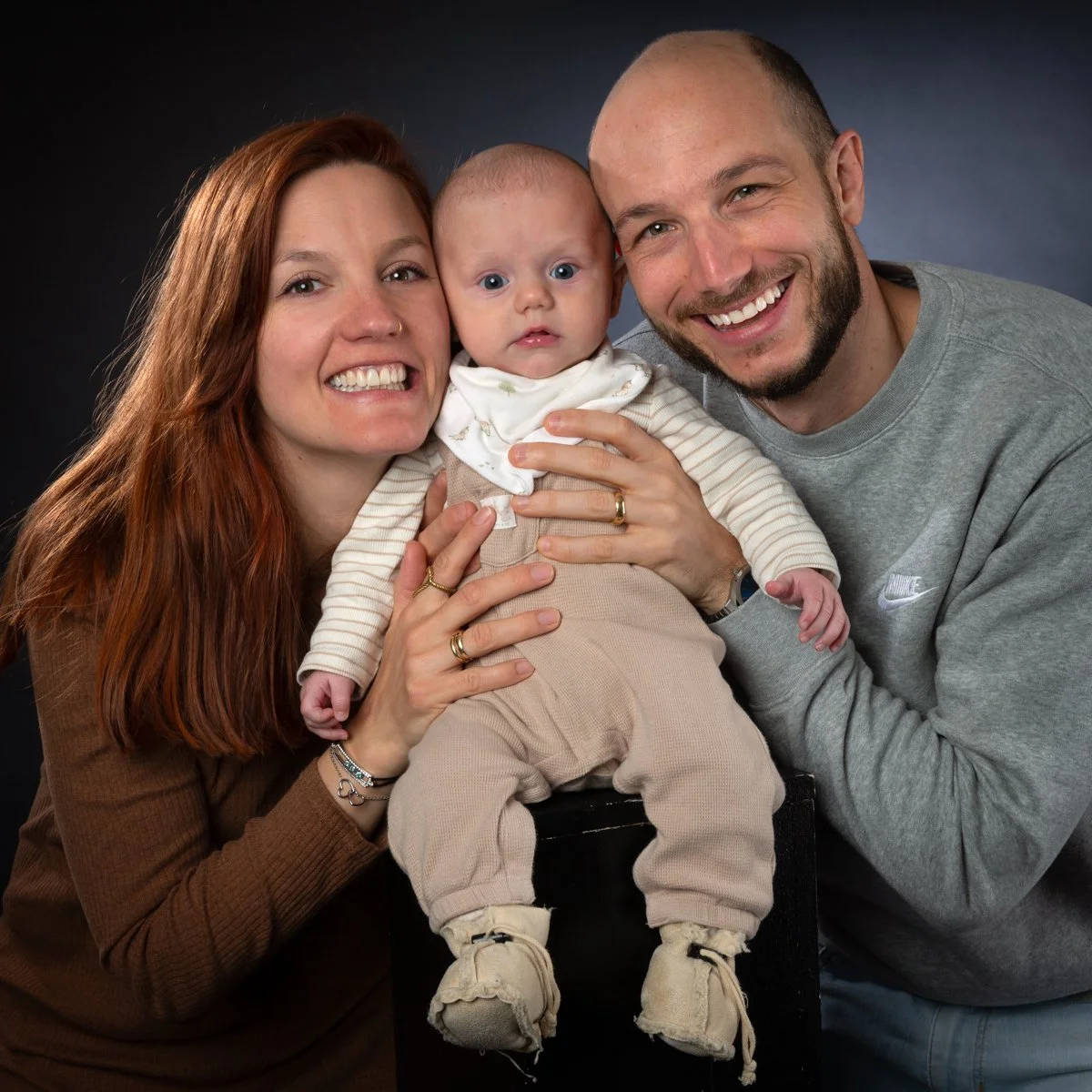 Une famille souriante composée d'une mère aux cheveux roux, d'un père chauve avec une barbe, et d'un bébé au centre, tous posant devant un fond sombre.