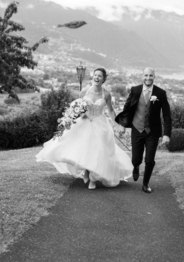 Un couple de mariés marchant main dans la main dans un parc avec un paysage de montagnes en arrière-plan, la mariée portant une robe de mariage et tenant un bouquet, sourire joyeux.