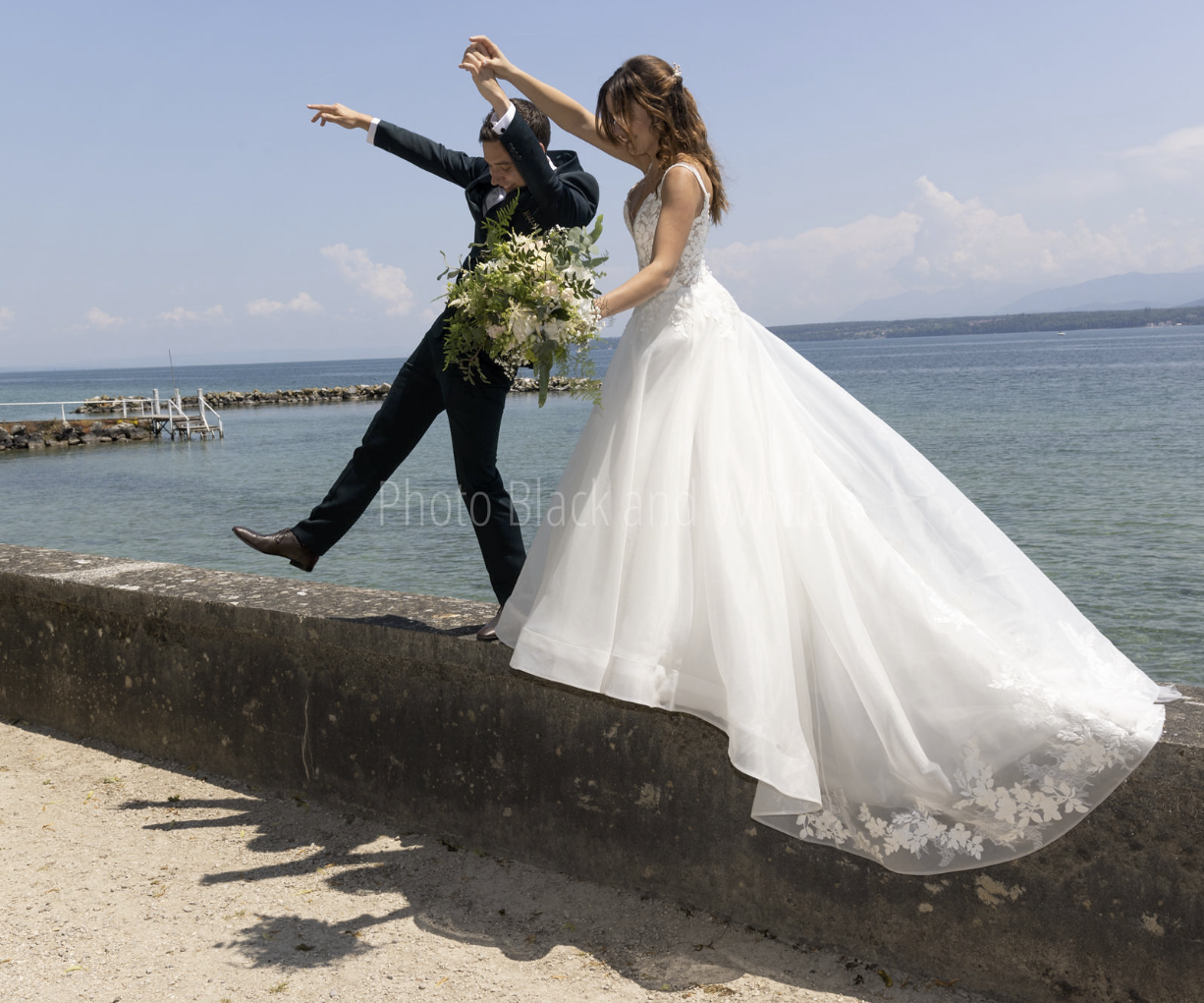 Un couple de mariés en vêtements de mariage, debout sur un mur en pierre au bord de la mer, avec le marié qui soulève la mariée. La mariée porte une robe de mariée blanche avec une longue traîne, et tient un bouquet de fleurs. La scène est en plein air avec un ciel ensoleillé et une mer calme en arrière-plan.