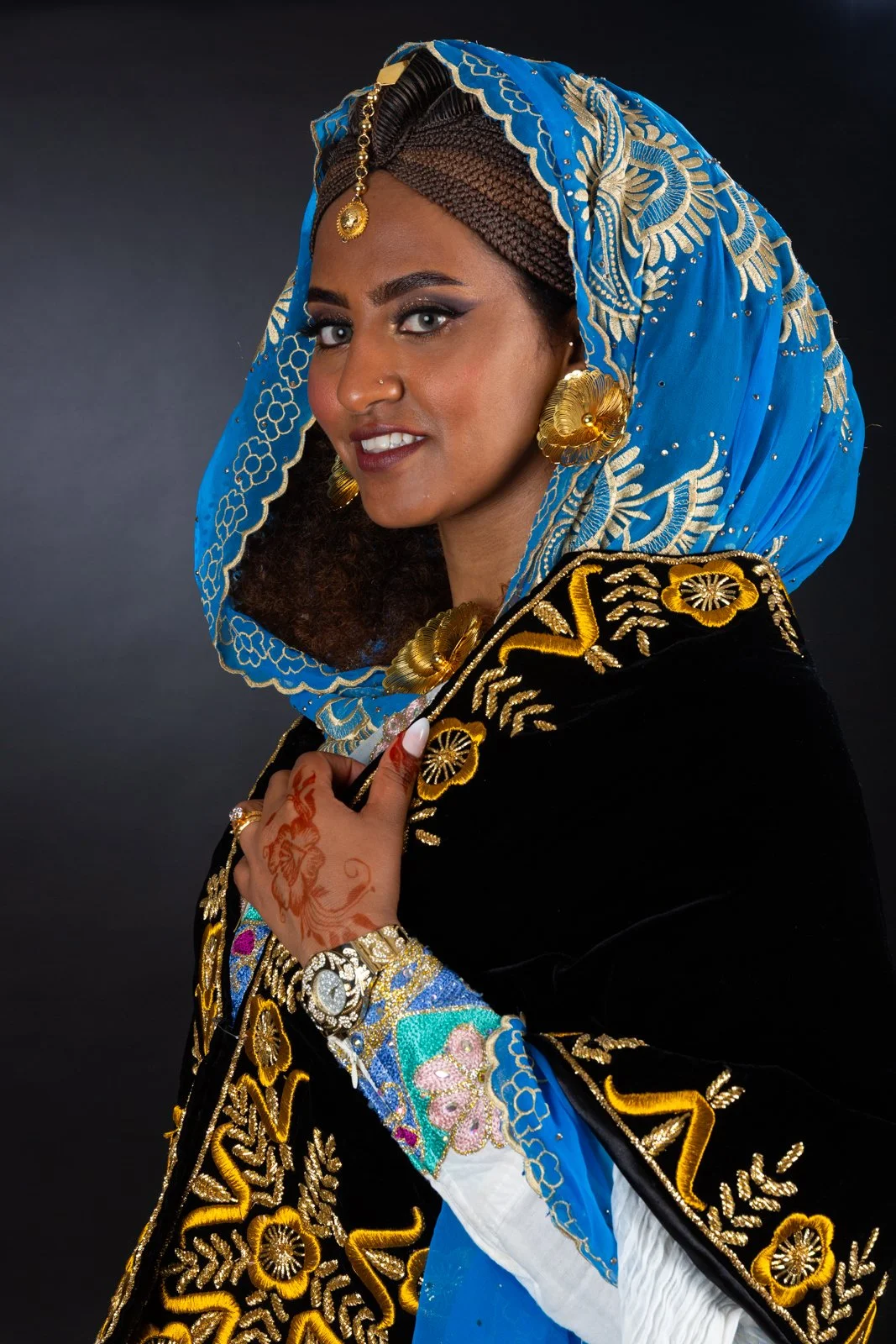Femme portant un foulard bleu et vêtements traditionnels noirs et dorés, avec des bijoux en or et des tatouages au henné, contre un fond sombre.
