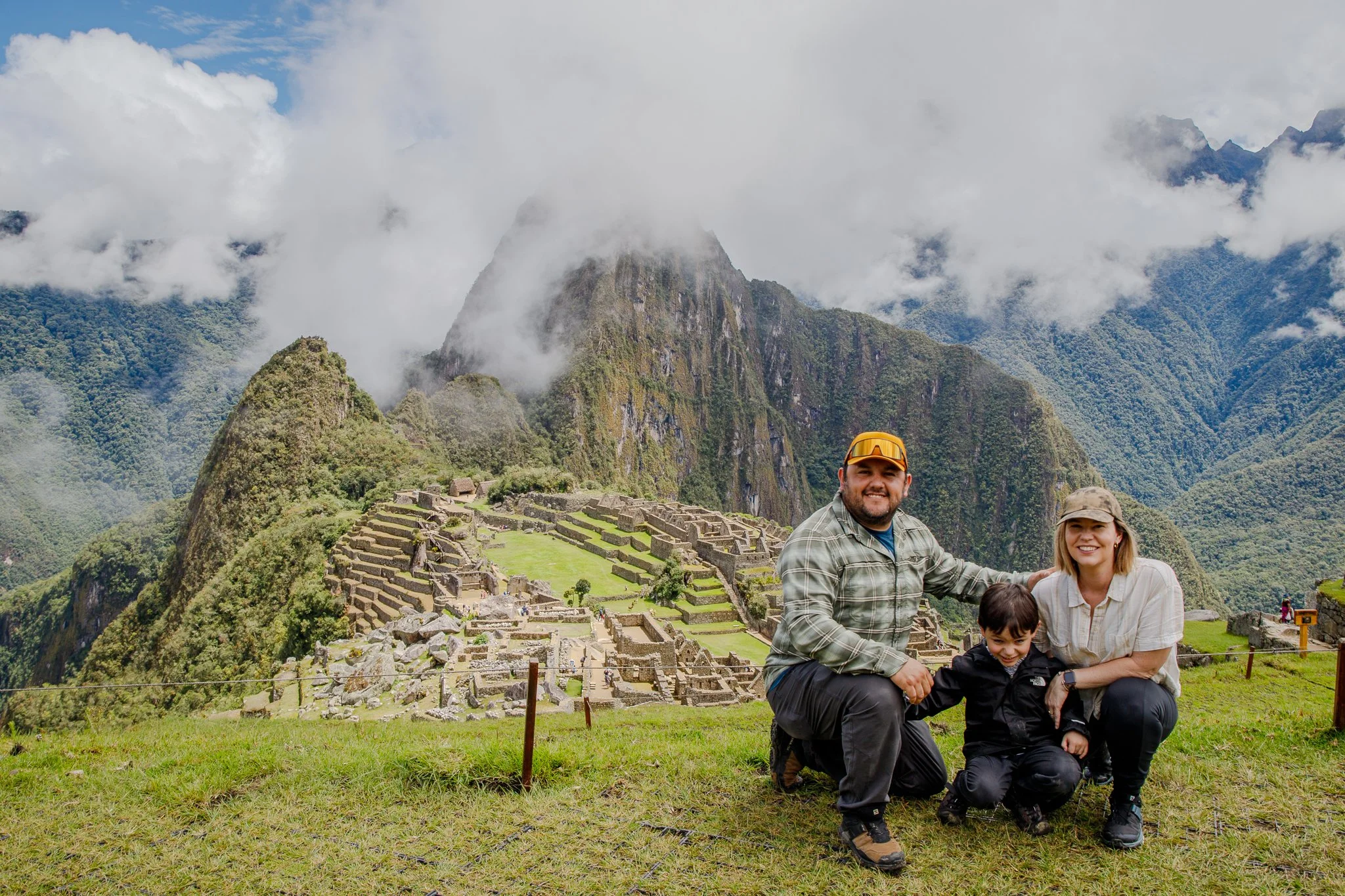 A family of four smiling in front of Machu Picchu with green mountains and cloudy sky in the background.