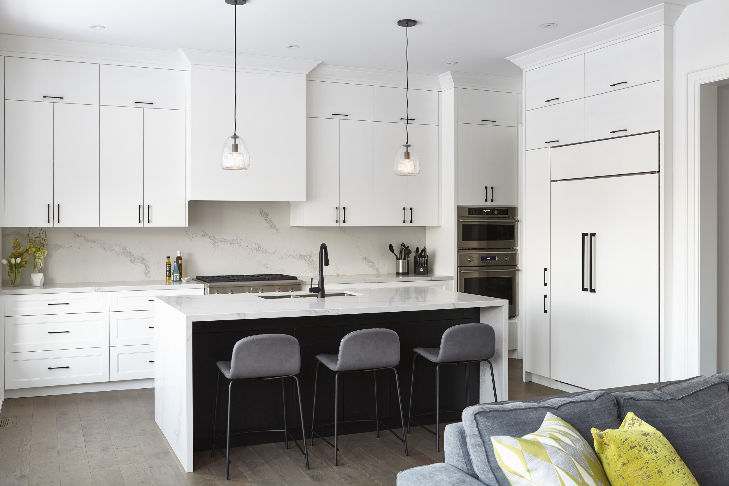 Modern kitchen with white cabinets, island with black sink, gray stools, pendant lights, stainless steel appliances, and a gray sofa with yellow pillows.