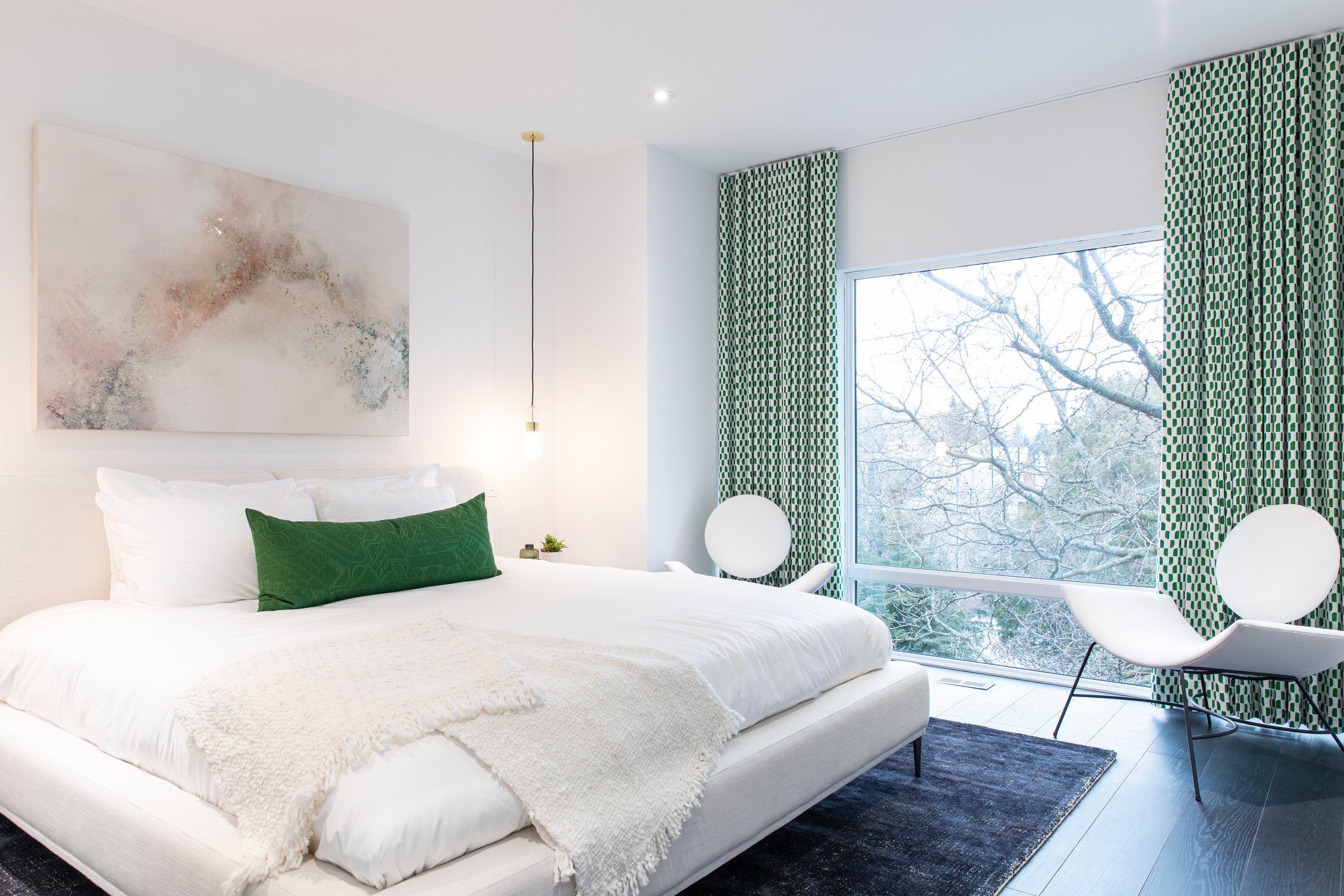 Modern bedroom interior with white bed, green accent pillow, abstract wall art, pendant light, large window, green patterned curtains, and two white chairs.