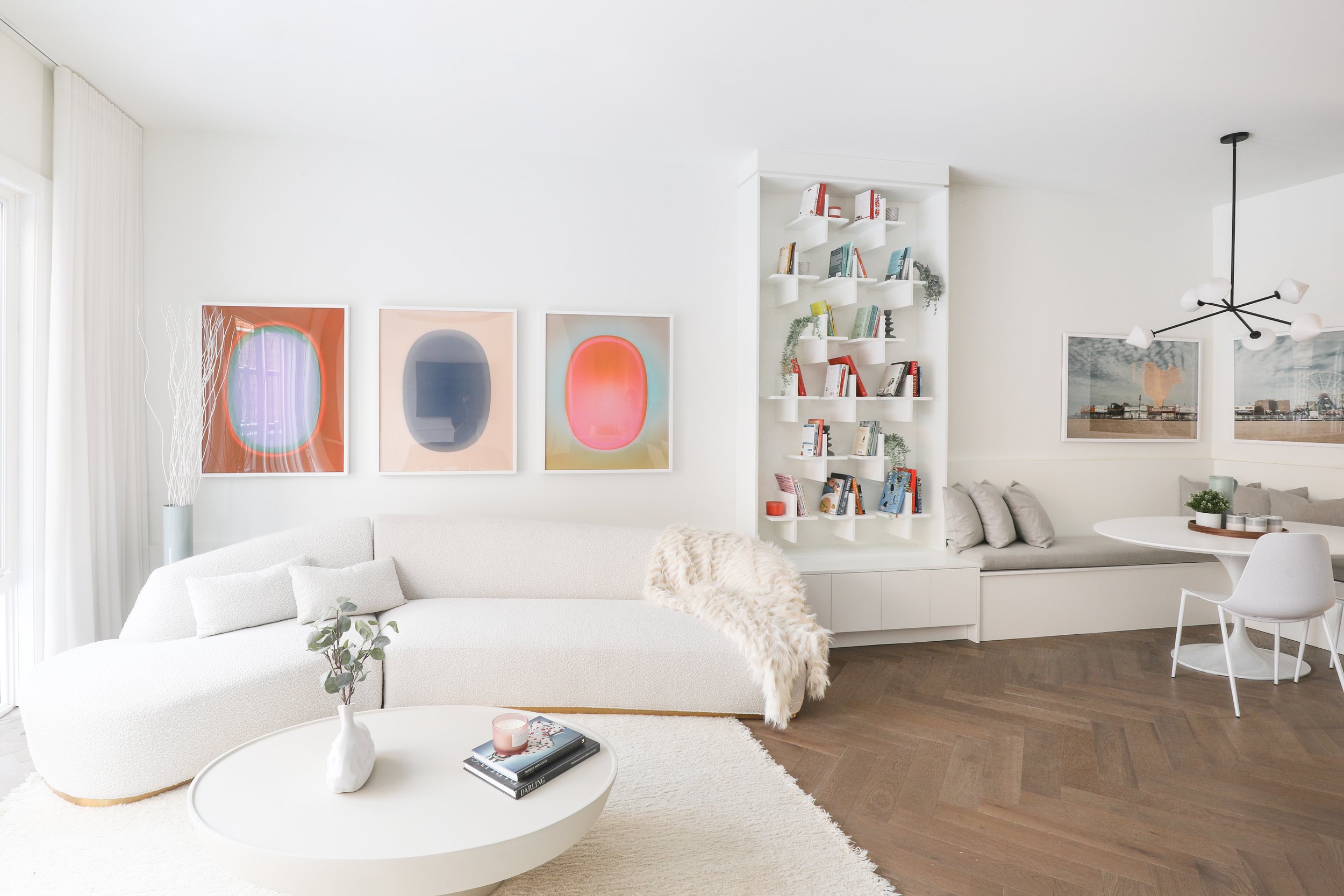 Modern living room with white curved sofa, round coffee table, abstract artwork, wall-mounted bookshelf, and dining nook with white table and chairs.