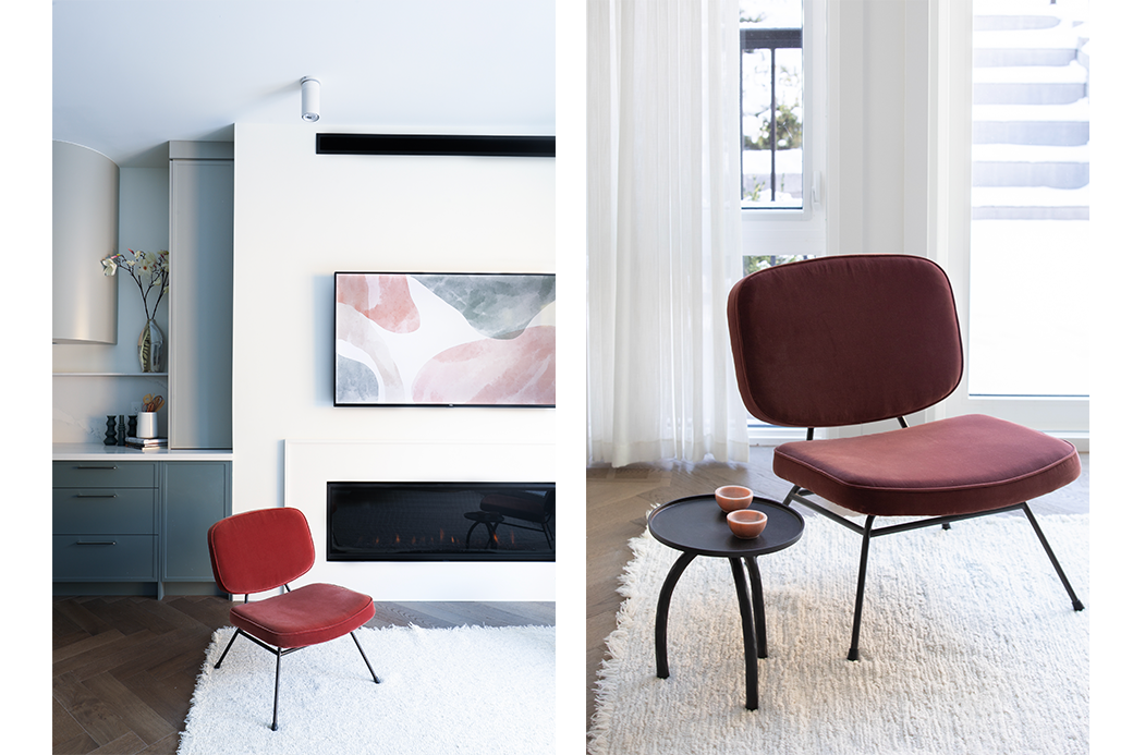 Modern living room featuring a red chair, a small round black side table, and a minimalist fireplace beneath a wall-mounted abstract painting. Light-filled room with sheer white curtains and a vase with flowers on a shelf.