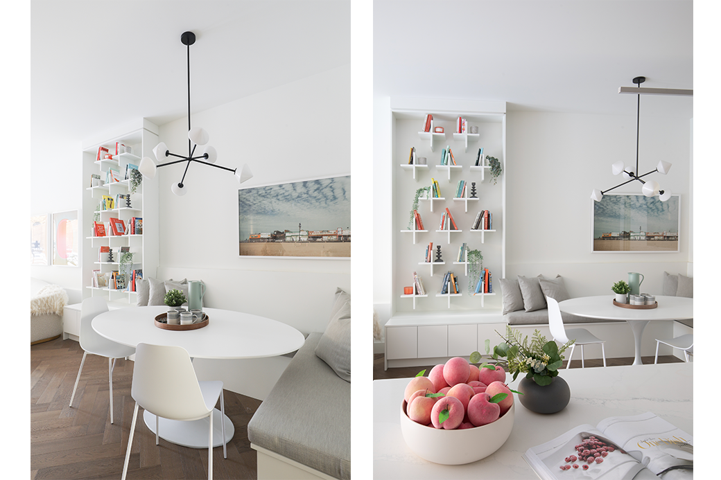 Modern dining area with white furniture, round table, contemporary chandelier, wall art, and built-in shelves filled with books and decor. A bowl of peaches and a small plant on the countertop.