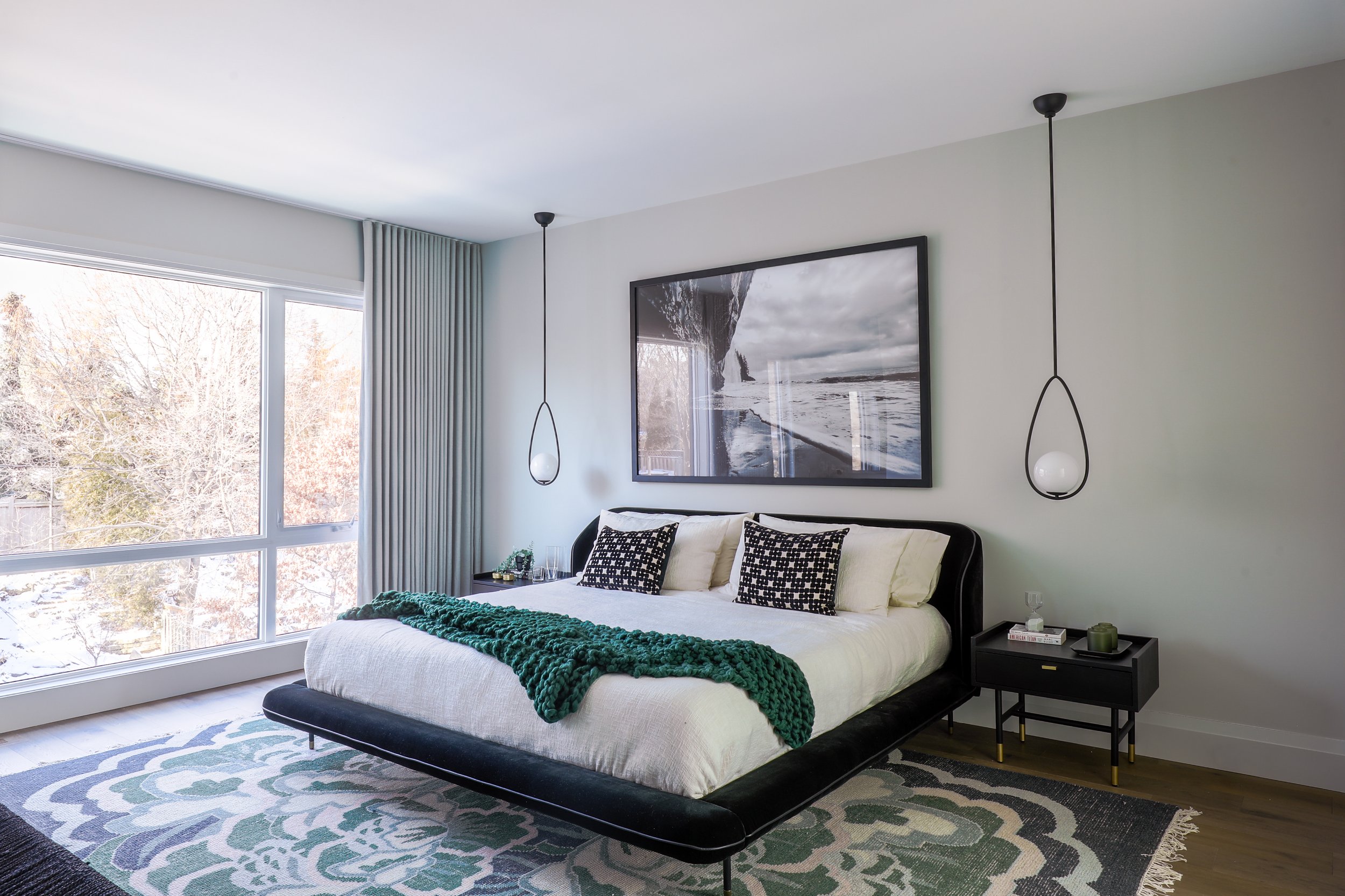 Modern bedroom with large window, abstract art, and pendant lights. Features a bed with white and black bedding, green throw, and black nightstand.