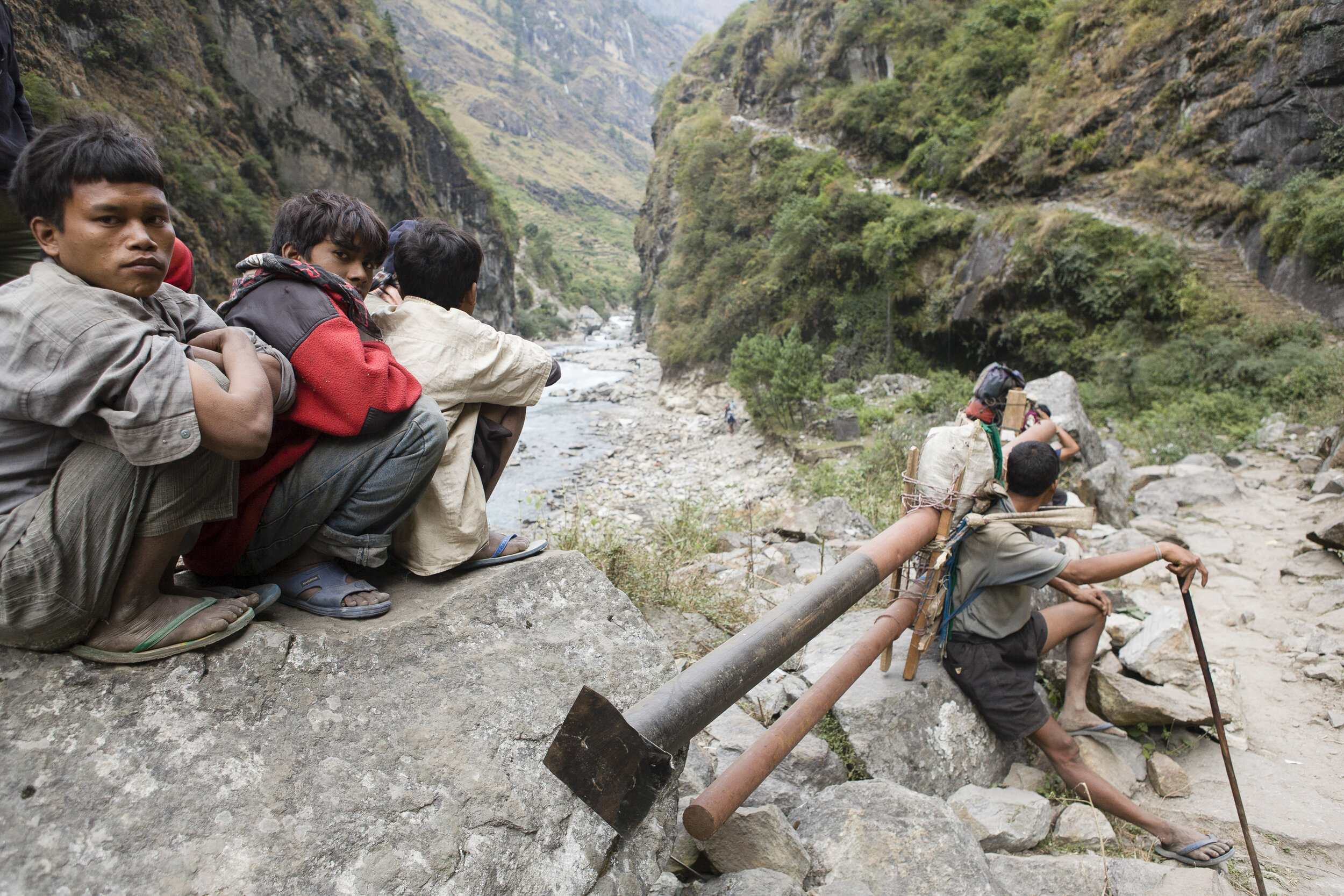  Nepali porters wait until workers dynamite rock along the Annapurna Circuit trail, near Tal. One carries telephone poles by bearing the weight on a strap placed on his forehead. Many porters wear plastic flip-flops while carrying heavy and awkward l