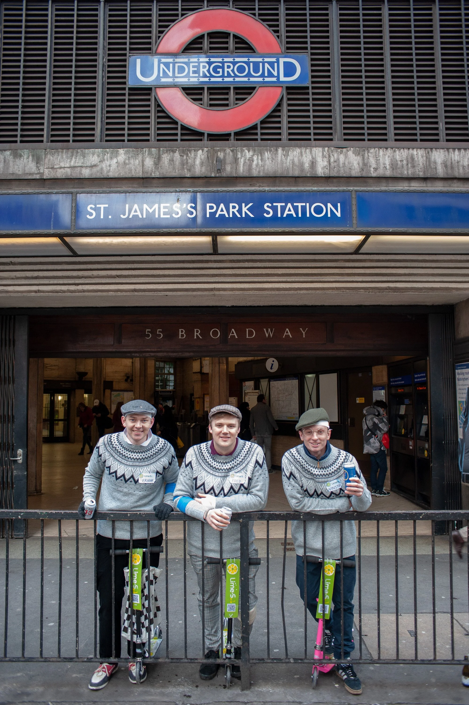 Three people in matching gray sweaters with white designs pose with scooters in front of the entrance to St. James's Park Station, London Underground.