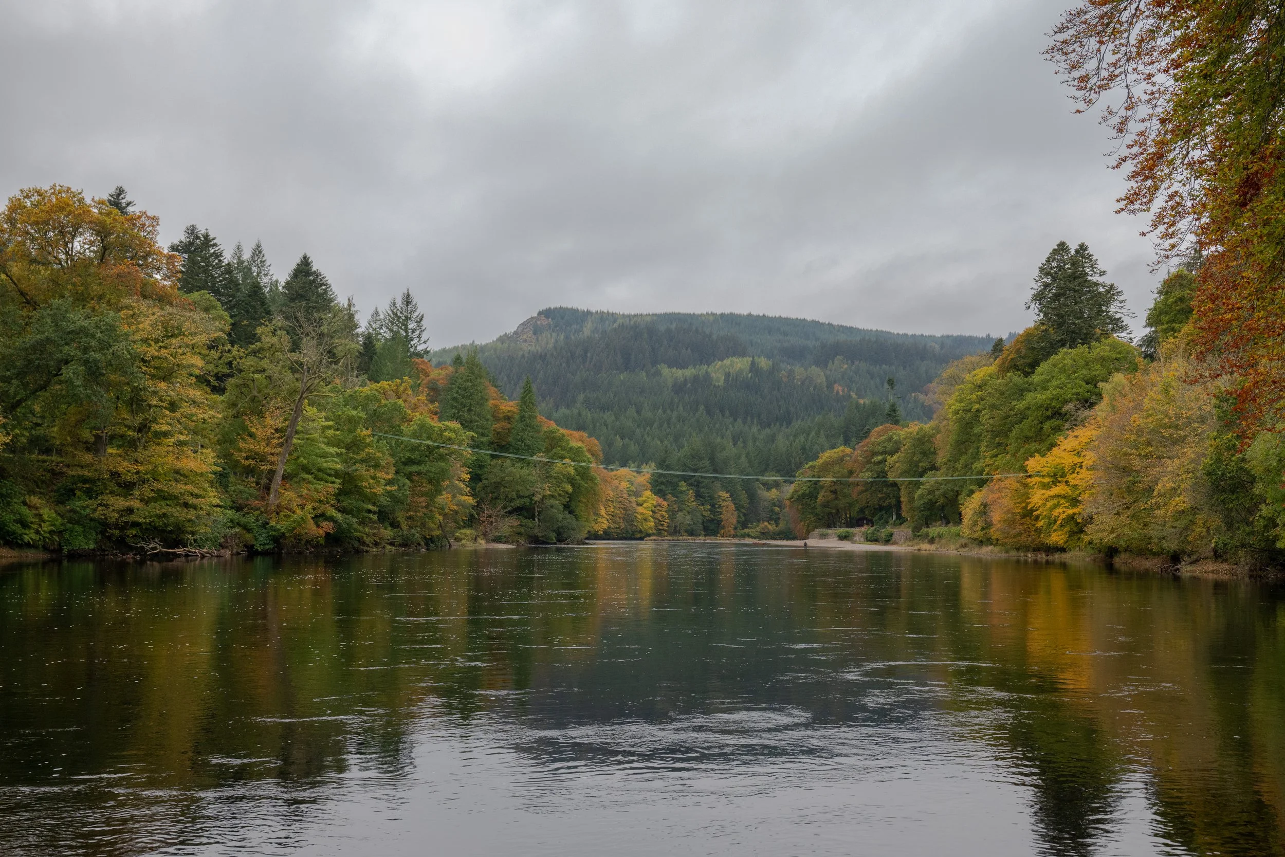 A wide shot of a calm river Tay reflecting trees with autumn colors, leading to a forested hill in the distance under a cloudy sky.