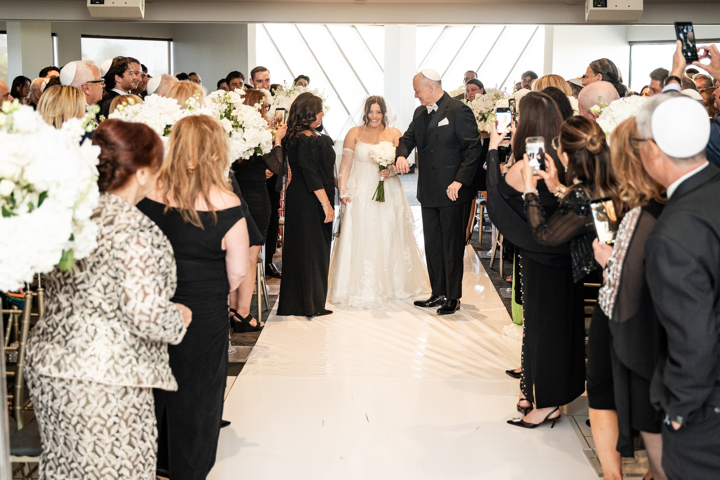 A wedding ceremony with a bride in a white dress and veil, holding a bouquet of white flowers, walking down the aisle flanked by guests taking photos and videos, with some guests wearing yarmulkes, in a modern indoor venue with large windows.