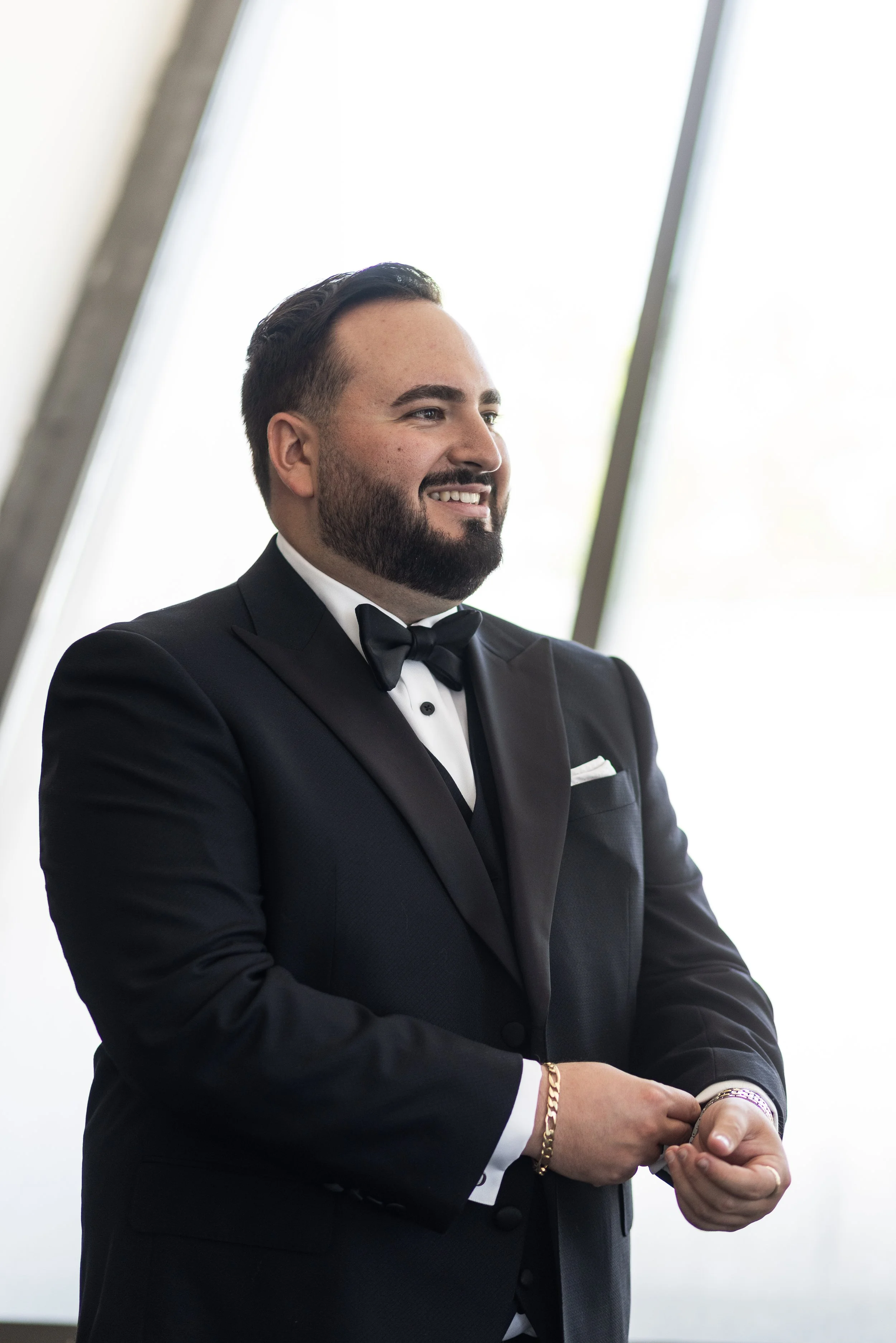 A man wearing a black tuxedo with a bow tie, smiling and looking away, standing indoors near a large window with bright natural light.