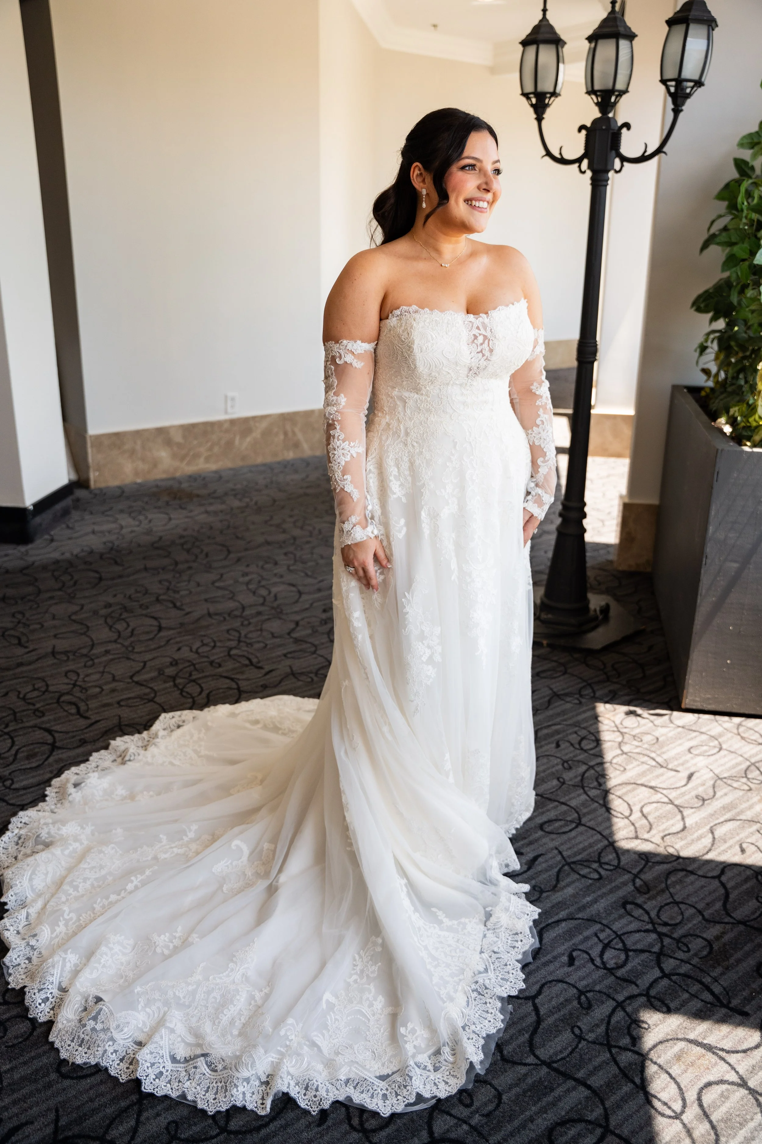 A woman in a wedding dress standing indoors beside a black antique-style lamp post and a potted plant, smiling and looking to her right.
