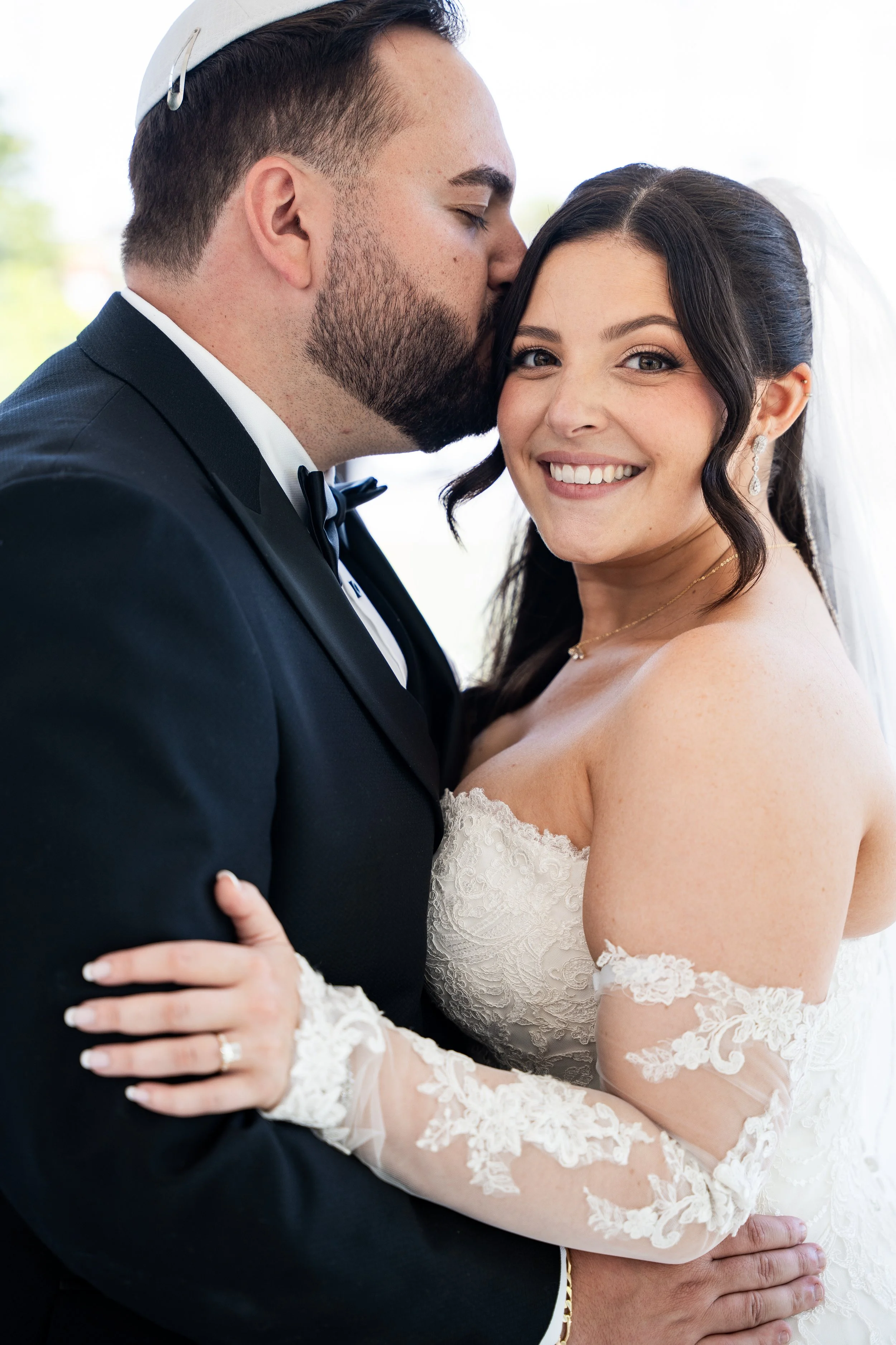 A newlywed couple embracing, with the groom kissing the bride on the forehead. The bride is smiling and wearing a wedding dress with lace details, and the groom is in a tuxedo with a bow tie.