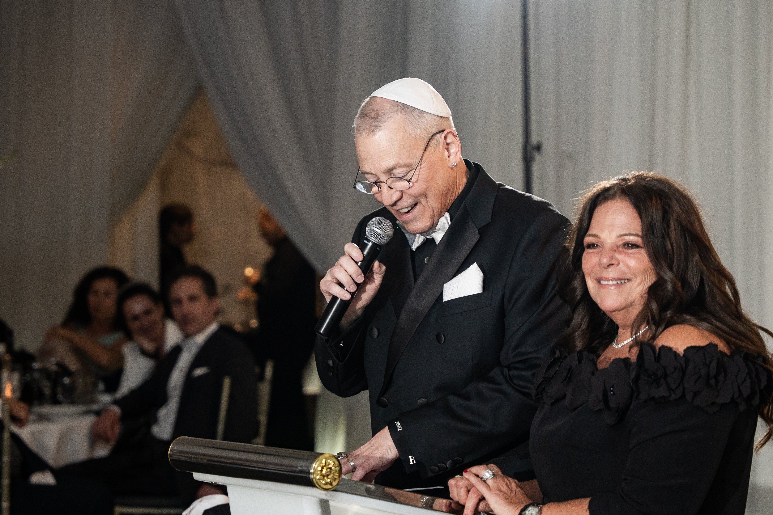An elderly man wearing glasses and a kippah, dressed in a tuxedo, speaking into a microphone at a formal event. Sitting beside him is a woman with long dark hair, wearing a black dress and pearl necklace, smiling. There are guests in formal attire se