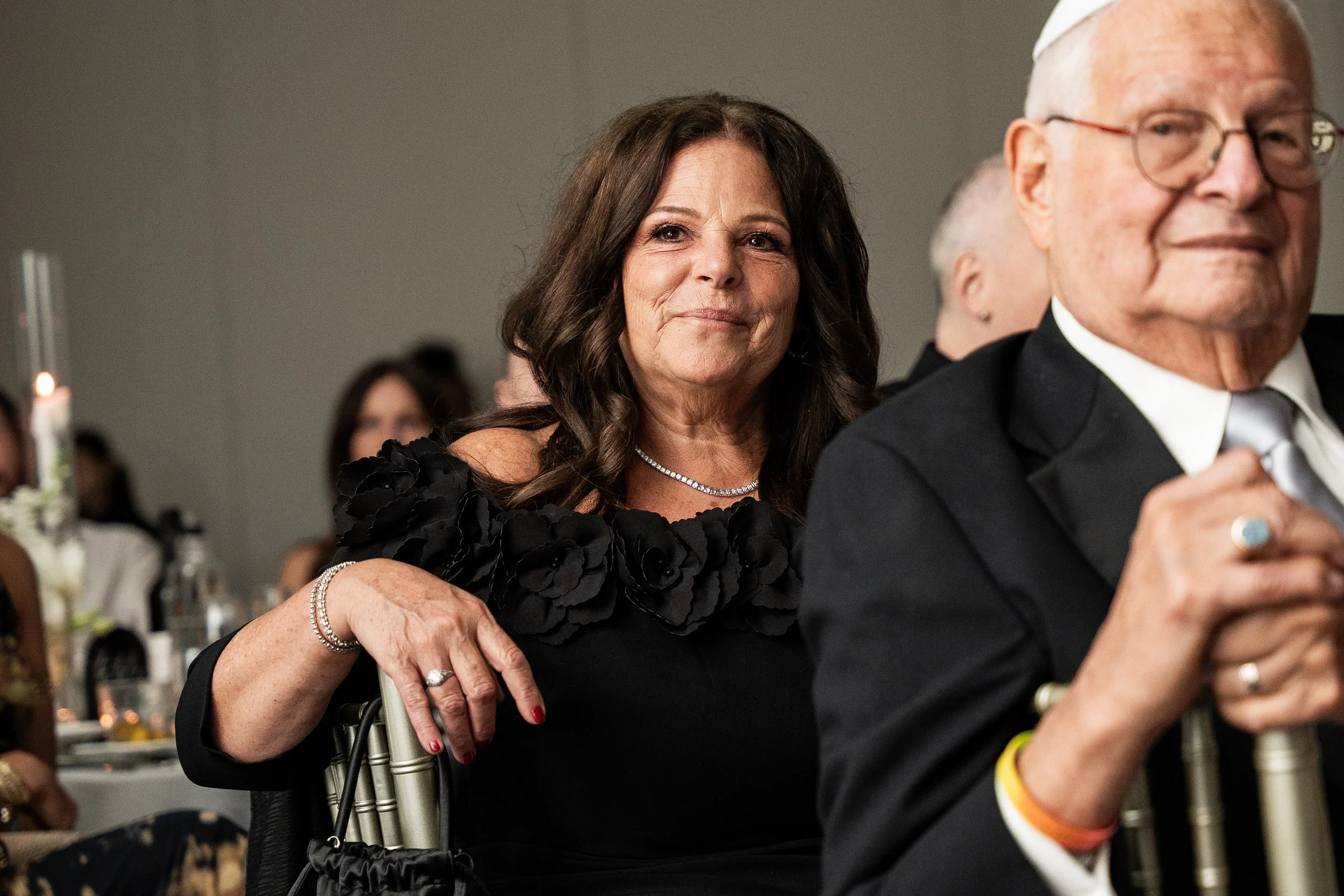 A woman with dark hair and a black dress, sitting next to an elderly man with glasses and a tuxedo, at a formal event. The woman has her hand on the man's arm and is smiling subtly.