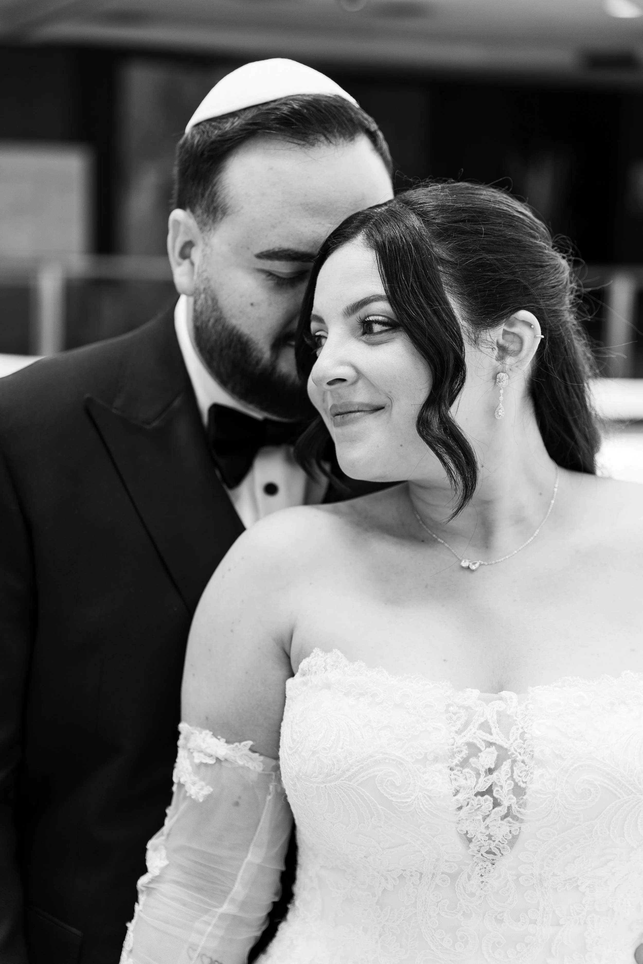 A black and white photo of a bride and groom. The groom is wearing a tuxedo with a bow tie and a yarmulke. The bride has dark hair styled in loose waves and is wearing elegant earrings and a strapless lace wedding gown. They are close, smiling and ga